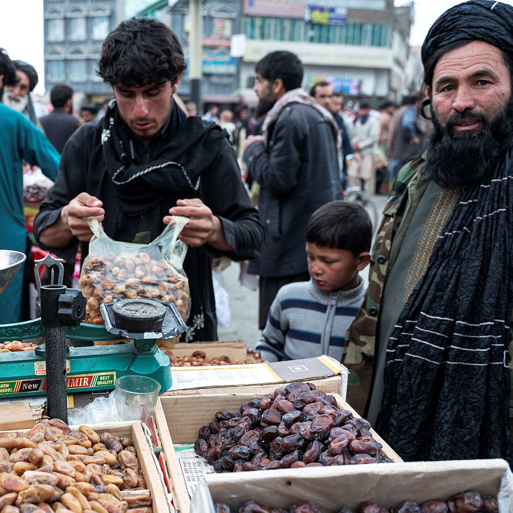 La imagen muestra un mercado al aire libre en un área concurrida. En el primer plano, hay un hombre de barba con un turbante que está de pie junto a una balanza y cajas llenas de dátiles. Otro hombre, que parece estar pesando los dátiles, está en el proceso de seleccionar los productos. También hay un niño pequeño cerca del hombre con la barba. El ambiente es activo, con varias personas en el fondo, lo que sugiere un bullicio típico de un mercado local. Las cajas de productos destacan por su variedad y colorido.