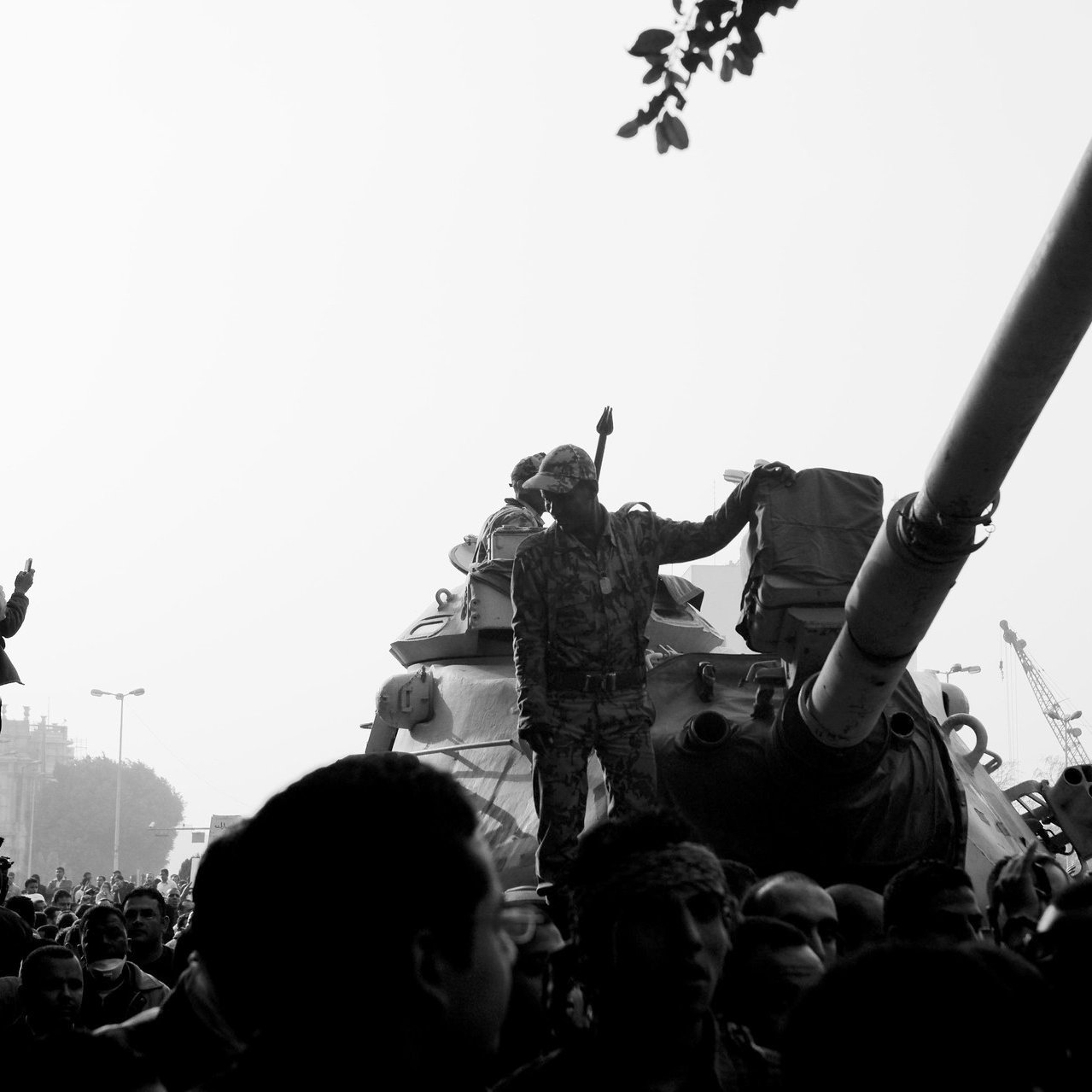 L'image montre une scène de manifestation avec un tank au premier plan. Des soldats sont visibles, dont un se tient debout sur le tank, levant un bras en signe de protestation ou de triomphe. Au premier plan, on aperçoit une foule de manifestants, certains prenant des photos ou filmant la scène. L'atmosphère est tendue, peut-être en raison de la situation politique. L'image est présentée en noir et blanc, ce qui renforce le côté dramatique de la scène.
