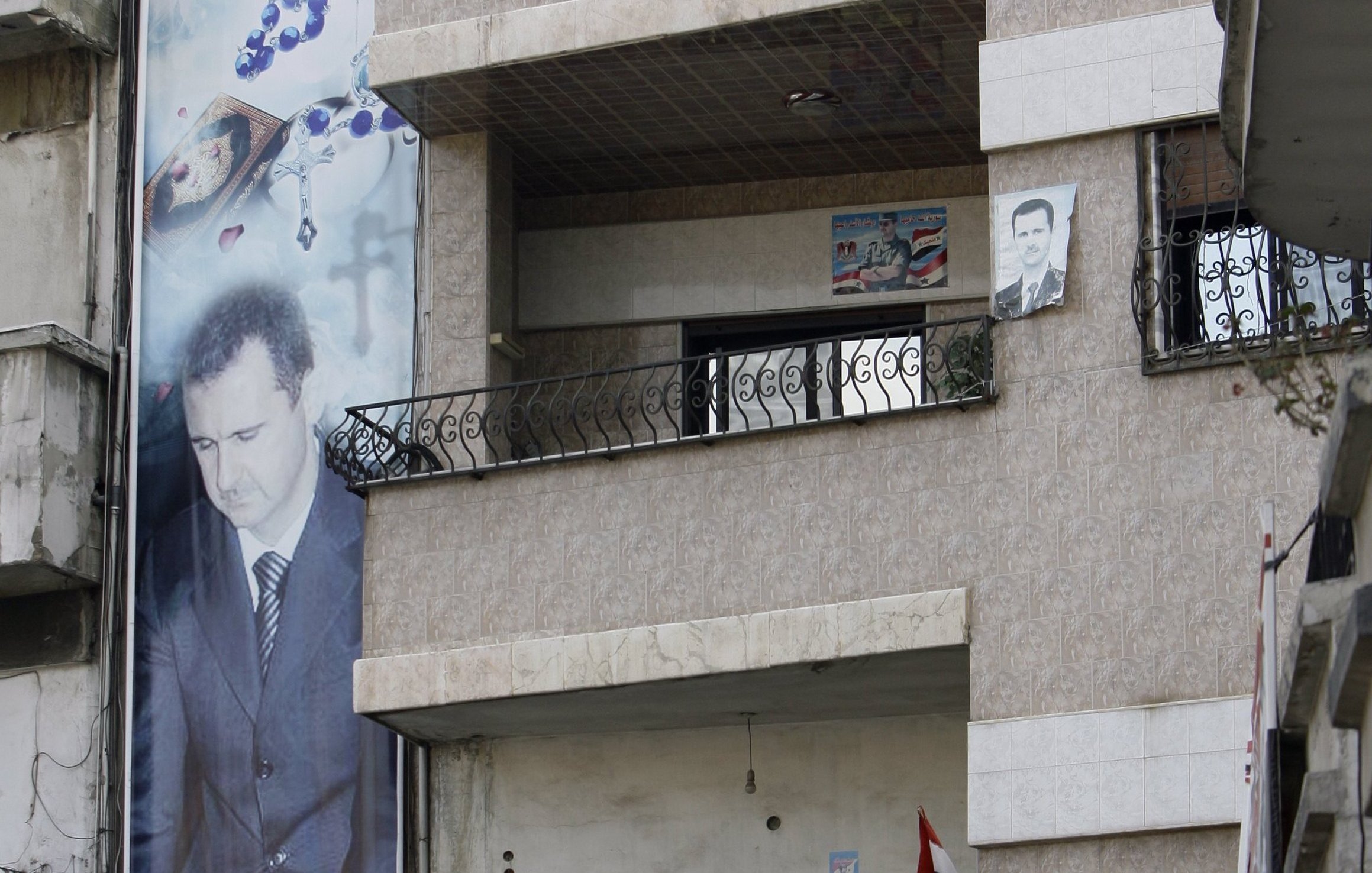 در این تصویر، نمایی از یک ساختمان چند طبقه دیده می‌شود که در آن پوسترها و تصاویر بزرگی از یک شخص (که احتمالاً یک شخصیت سیاسی است) نصب شده است. دو مرد در بالکن طبقه دوم ایستاده‌اند و به فضای بیرون نگاه می‌کنند. همچنین، در زیر ساختمان، بنرهایی با شعارها و تصاویری از دیگر افراد نصب شده است. محیط به نظر می‌رسد که در یک منطقه شهری باشد و برخی نشانه‌های ملی مانند پرچم در تصویر دیده می‌شود.