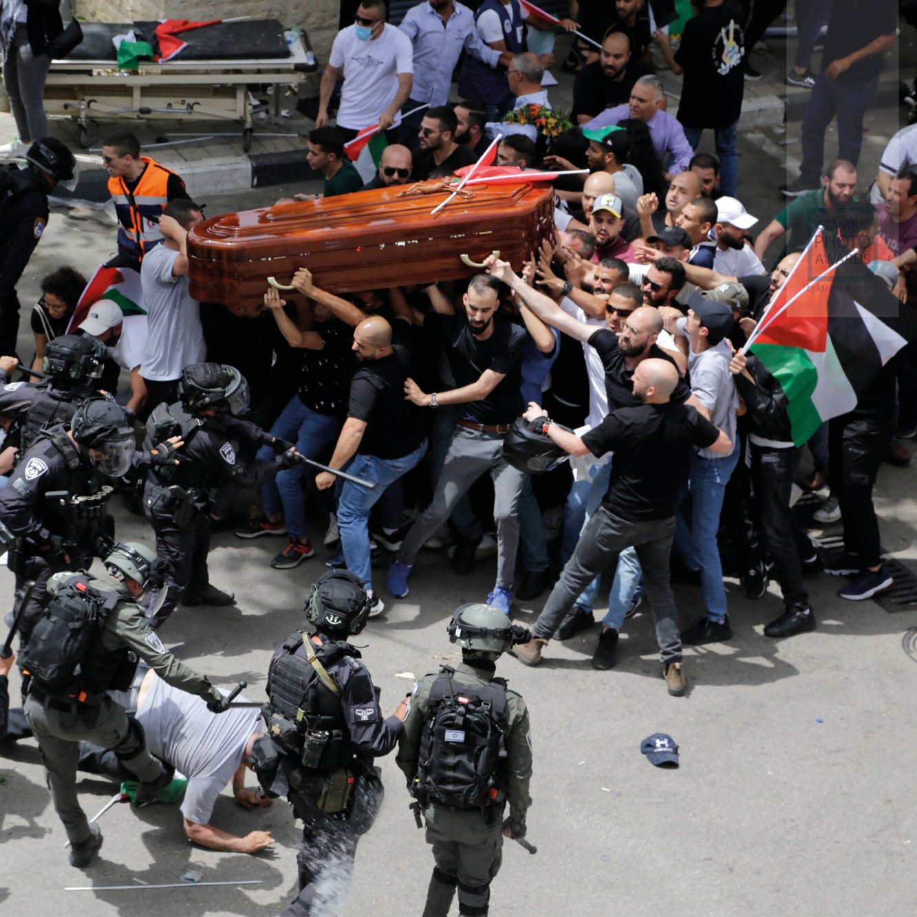 A crowd confronts police while carrying a coffin; flags and tension are visible.