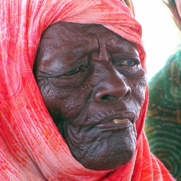 L'image montre un portrait d'une personne âgée avec une expression réfléchie. Elle porte un foulard coloré, qui semble être enroulé autour de sa tête, et son visage présente des rides marquées, témoignant de son âge et de son vécu. La lumière met en valeur les détails de son visage.