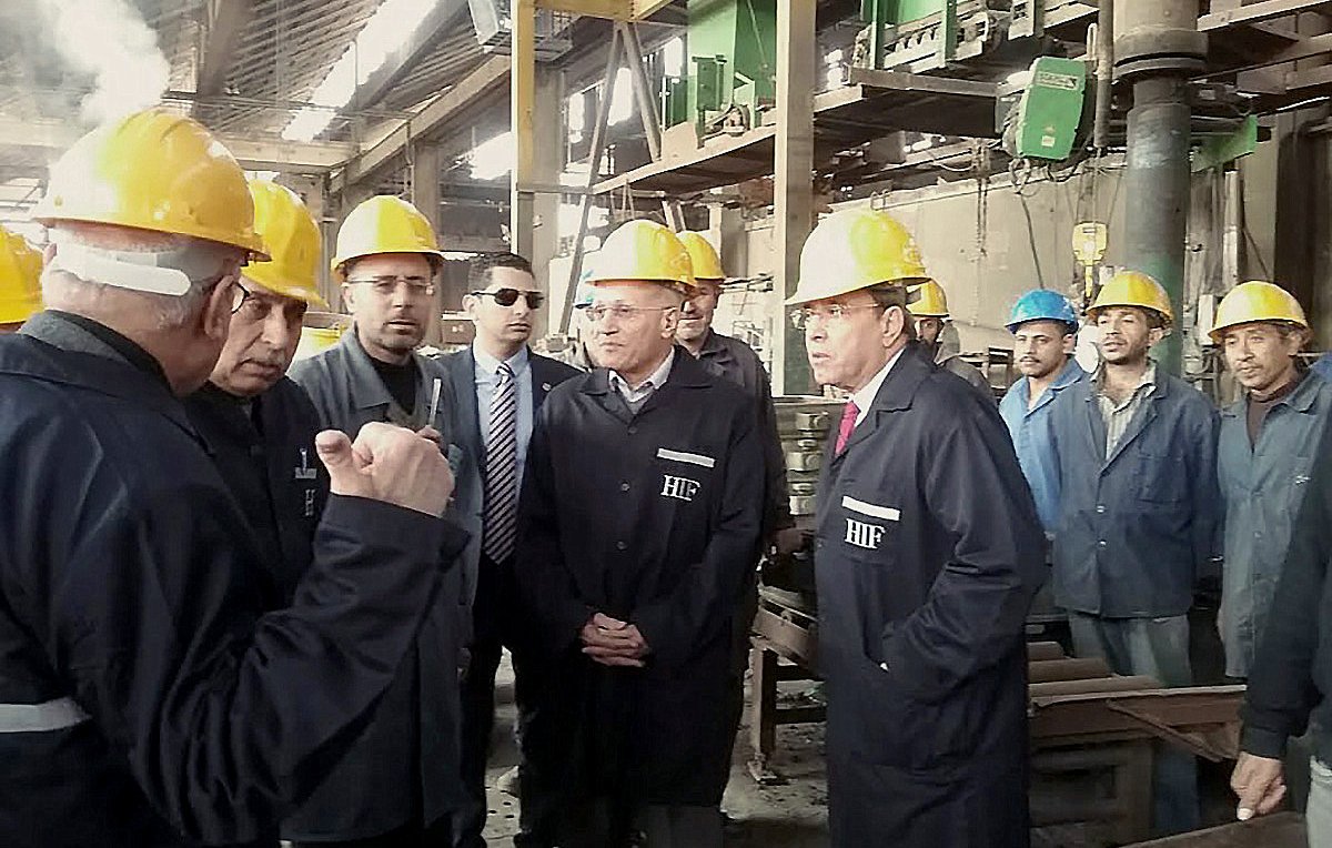 L'image montre un groupe de personnes dans un environnement industriel, probablement dans une usine. Les participants portent des casques de sécurité jaunes, ce qui indique qu'ils sont dans un lieu de travail où des précautions de sécurité sont nécessaires. Certains hommes sont en train de discuter ou d'écouter attentivement, tandis que d'autres observent la scène. En arrière-plan, on peut voir des machines et des équipements typiques d'un environnement de production. L'atmosphère semble être sérieuse et professionnelle.