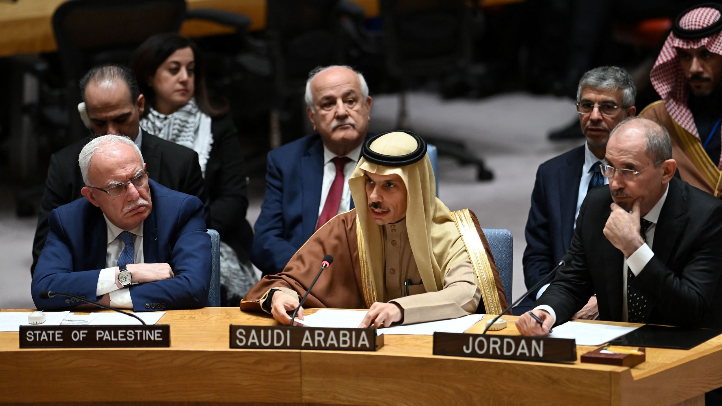 L'image montre une réunion de l'Assemblée générale ou du Conseil de sécurité de l'ONU. Plusieurs représentants de différents pays sont assis autour d'une table. À gauche, on voit un homme portant des lunettes avec un badge indiquant "État de Palestine". Au centre, un homme en tenue traditionnelle saoudienne prend la parole, tandis qu'un autre à droite, en costume noir, représente la Jordanie. En arrière-plan, d'autres participants écoutent attentivement. L'atmosphère semble sérieuse, suggérant une discussion importante.