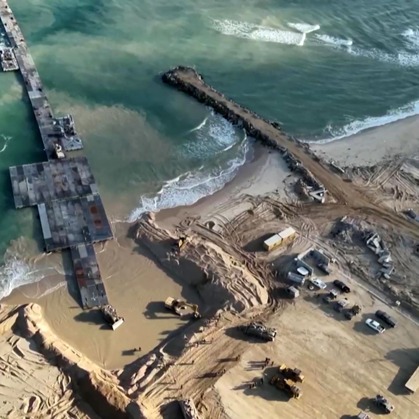 L'immagine mostra una vista aerea di una zona costiera in fase di costruzione o ristrutturazione. Si possono vedere macchine da lavoro e attrezzature presenti sulla spiaggia e nelle vicinanze, mentre un molo si estende verso il mare. Le onde del mare si infrangono sulla riva, e il terreno circostante appare movimentato e scavato, con evidenti segni di lavori in corso. La luce naturale esalta i dettagli del paesaggio, creando un contrasto tra l'acqua blu e la sabbia.