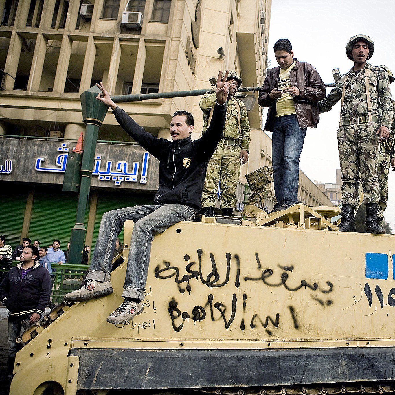 در این تصویر، یک گروه از مردان و سربازان در حال تجمع روی یک خودرو نظامی هستند. چند نفر از آنها به سمت دوربین خوشحالی می‌کنند و دست بلند کرده‌اند. در پس‌زمینه، ساختمان‌هایی با نشانه‌ها و نوشته‌هایی به زبان عربی دیده می‌شود. فضای تصویر نشان‌دهنده یک وضعیت اجتماعی یا سیاسی است که در آن افراد در حال ابراز احساسات و تعامل با یکدیگر هستند.