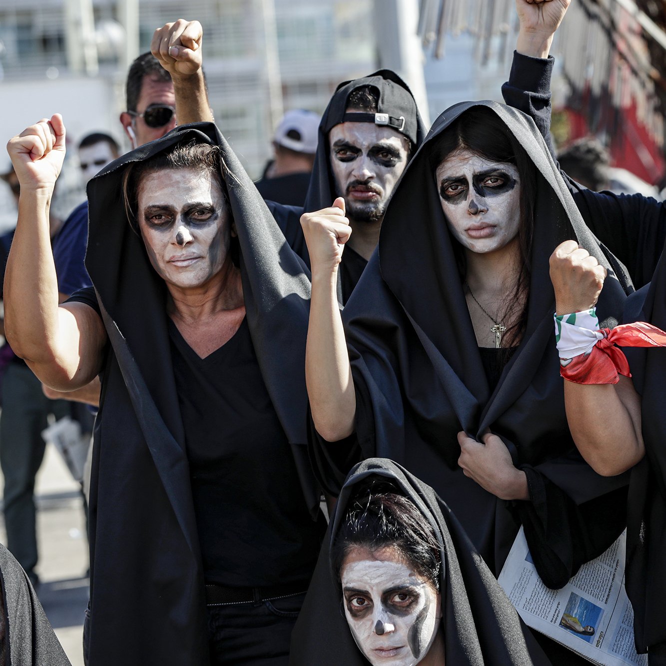 در این تصویر، گروهی از افراد زن و مرد را مشاهده می‌کنیم که لباس‌های سیاهی به تن دارند و صورت‌های خود را با رنگ سفید نقاشی کرده‌اند. آن‌ها در حال نمایش اعتراض خود هستند و برخی از آن‌ها با دستان بلند شده، نشانه‌ای از قدرت و اتحاد را نشان می‌دهند. پس‌زمینه تصویر به وضوح حاکی از یک تجمع یا تظاهرات است. حالت چهره‌ها جدی و معنادار به نظر می‌رسد، که بیانگر عزم و اراده آن‌ها برای بیان دیدگاه‌ها و خواسته‌هایشان است.