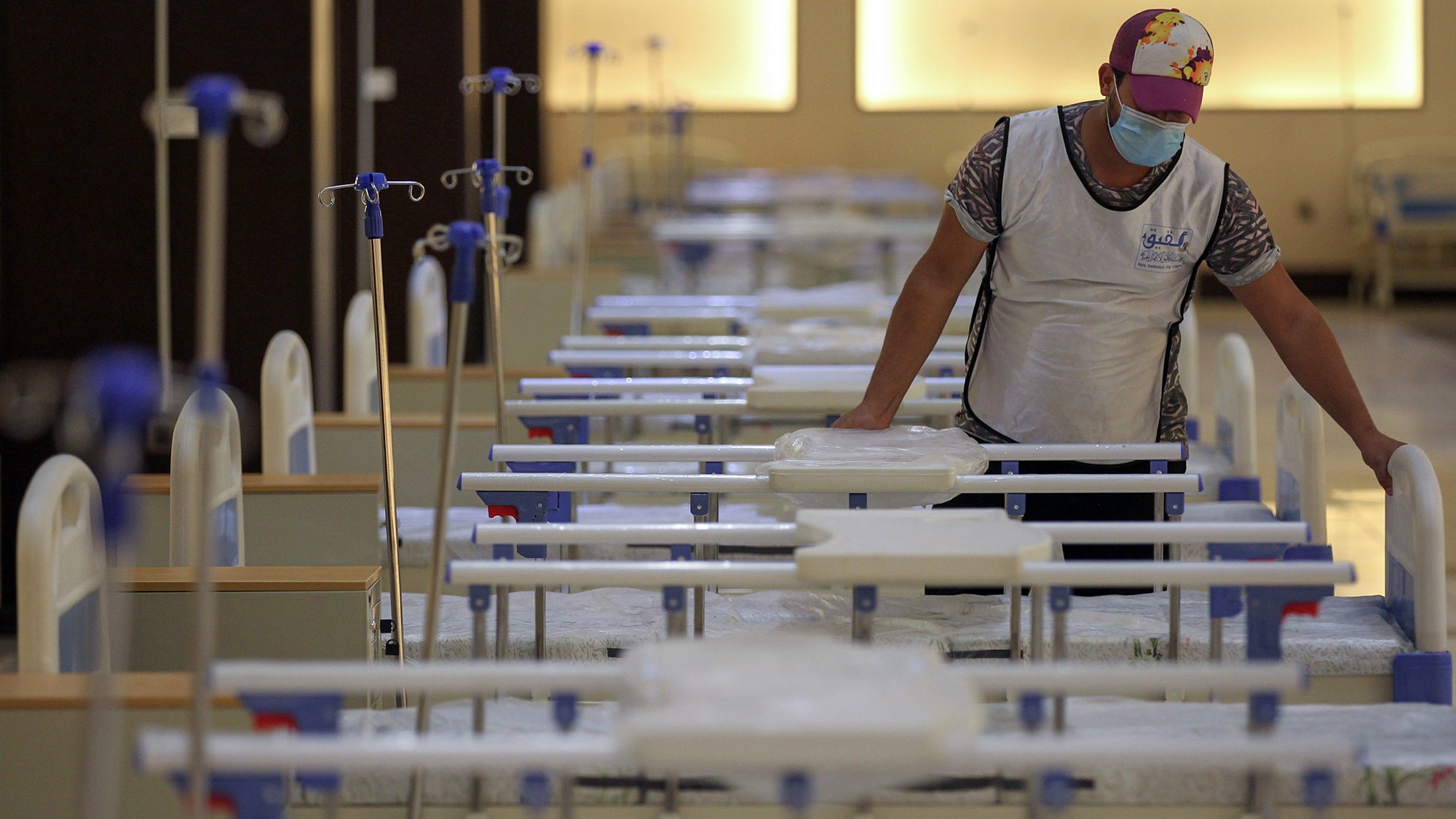 The image shows a person in a mask and a cap standing in a room filled with hospital beds. The beds are arranged in neat rows, and the area appears organized and prepared, possibly for medical purposes. The individual is interacting with the beds, which may indicate they are setting up or maintaining the space. The lighting in the room is warm and soft, contributing to a calm atmosphere.