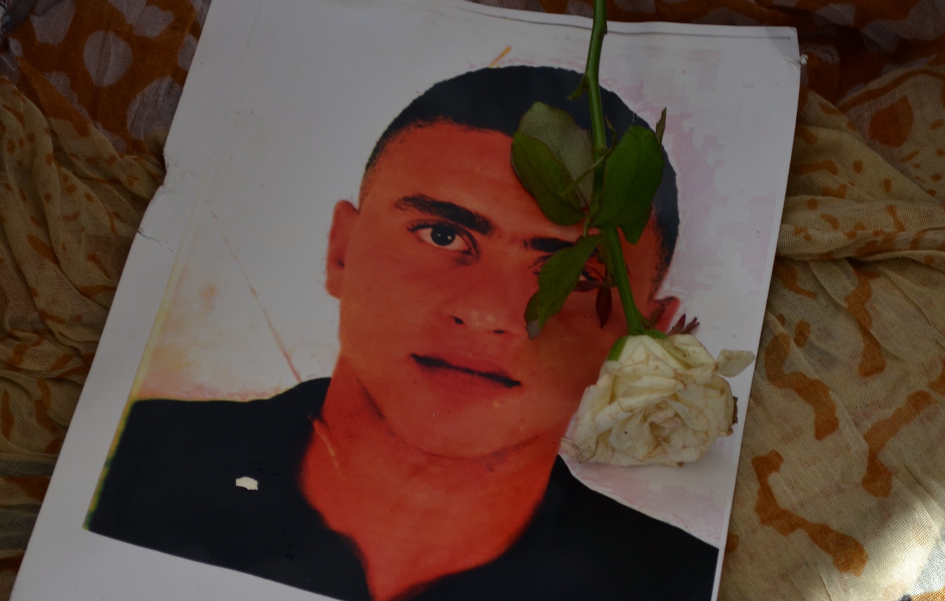 L'immagine mostra un ritratto di un giovane uomo posato su un tessuto colorato. Sopra il ritratto è adagiata una rosa bianca, che aggiunge un tocco di dolcezza e commemorazione. Il contrasto tra il volto del ragazzo e il fondo morbido e multicolore crea un'atmosfera di riflessione e rispetto.