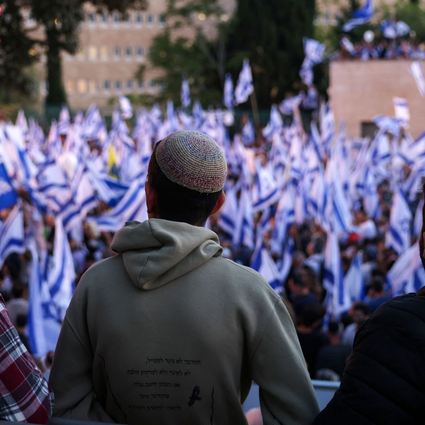 The image shows three individuals, viewed from behind, observing a large crowd at an outdoor event. The crowd is holding blue and white flags, which may symbolize a national identity. The individuals are wearing kippahs (traditional Jewish head coverings). The setting appears to be a public gathering, possibly a rally or demonstration, with trees and buildings visible in the background. The atmosphere seems lively and engaged.