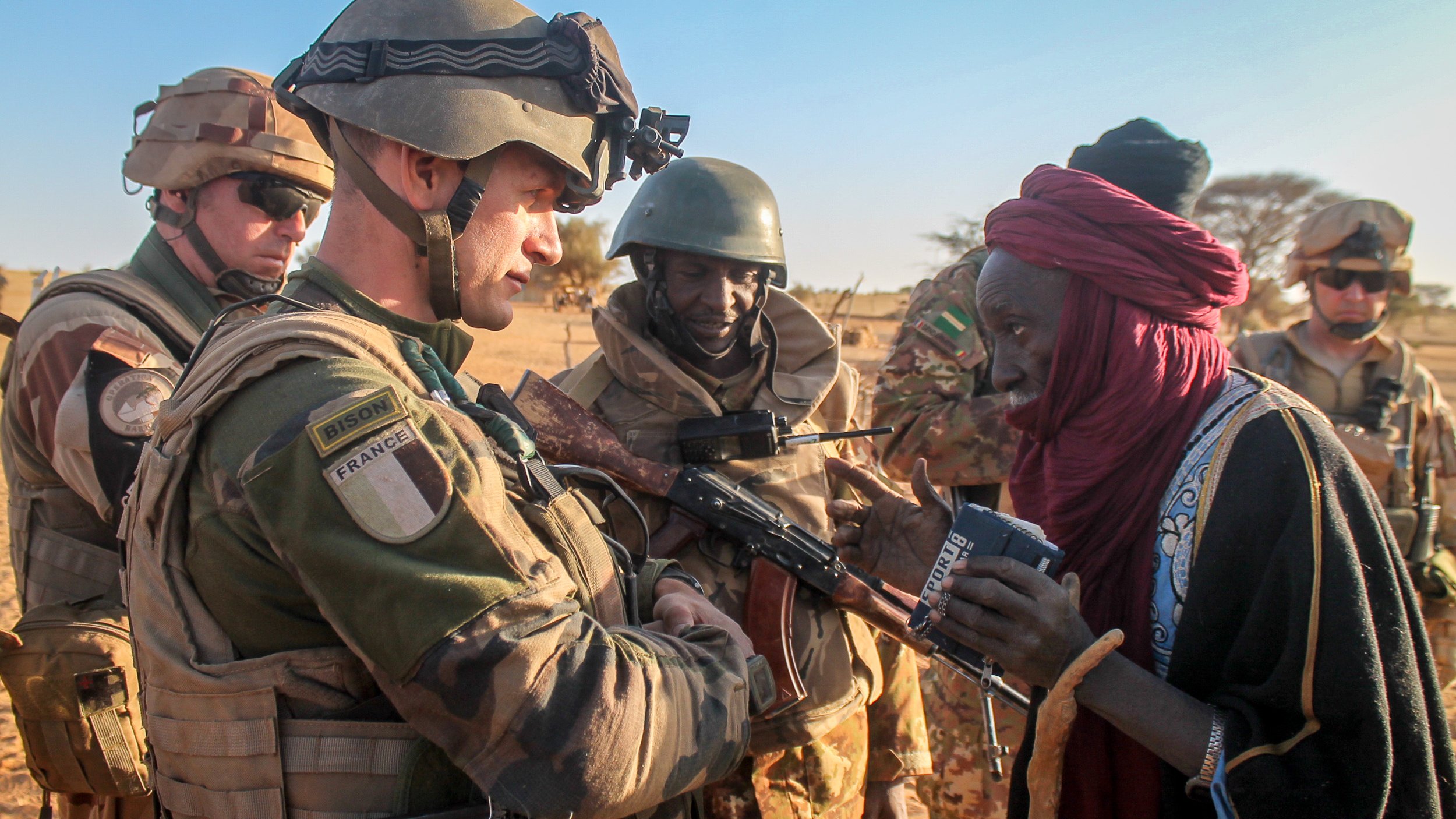 L'image montre un soldat en uniforme militaire, probablement français, interagissant avec un homme âgé, qui semble local. Le soldat est armé et porte un gilet pare-balles ainsi qu'un casque, tandis que l'homme a des vêtements traditionnels. En arrière-plan, d'autres militaires, également en uniforme, observent la scène. Le cadre semble désertique, typique d'une région saharienne, avec un ciel dégagé et un sol sablonneux. L'interaction semble empreinte de respect et d'échange culturel.