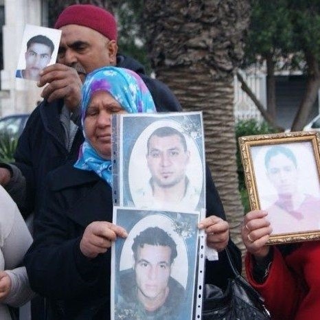 Nell'immagine si vede un gruppo di persone che partecipano a una manifestazione. Alcuni di loro tengono in mano delle fotografie di uomini. Le donne indossano il velo e mostrano espressioni serie, mentre gli uomini dietro di loro sembrano sostenere le stesse fotografie. L'ambiente intorno è urbano, con alberi e edifici visibili sullo sfondo. La scena trasmette un senso di protesta e richiesta di giustizia.