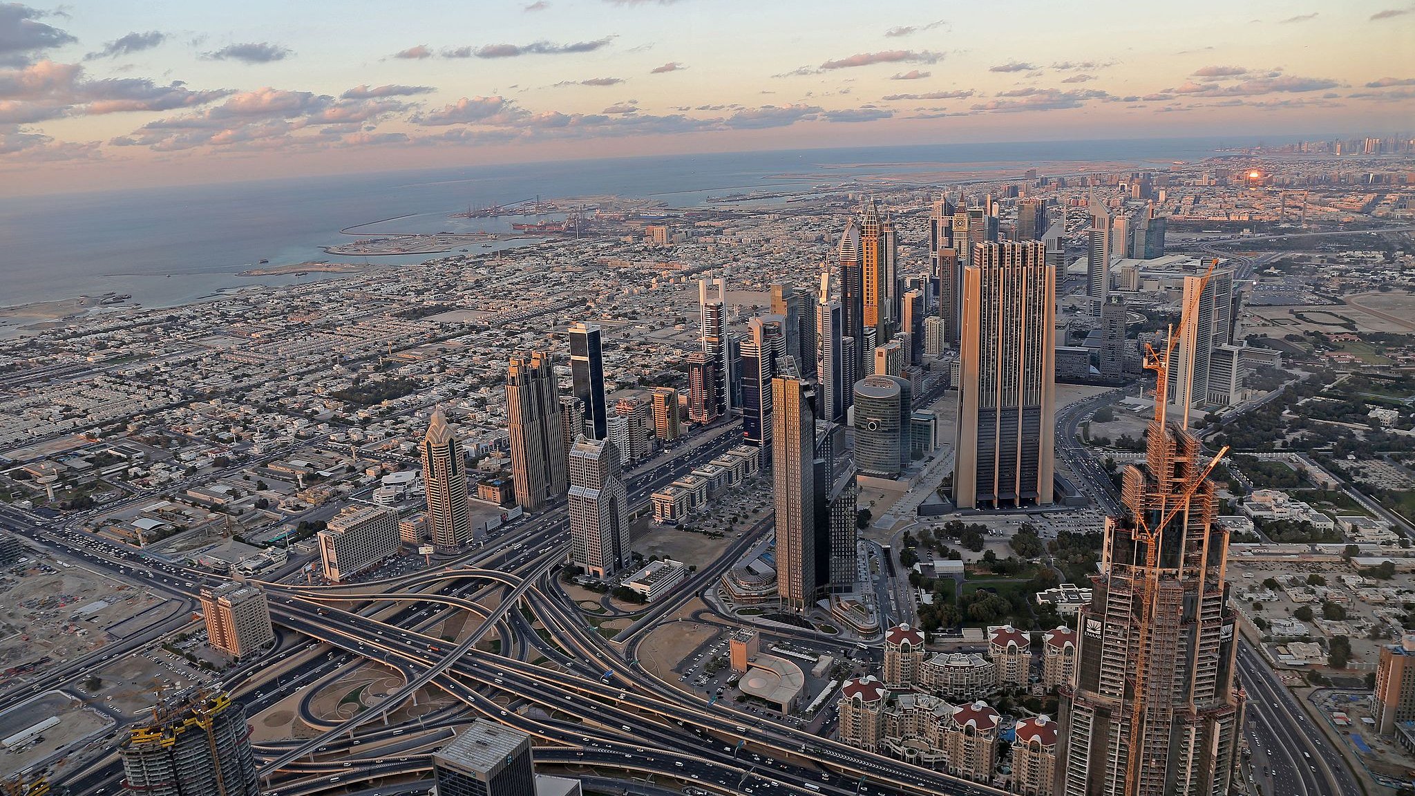The image depicts a stunning aerial view of a sprawling cityscape, likely a metropolitan area with modern architecture. Skyscrapers dominate the skyline, showcasing a mix of designs and heights. Below, intricate road systems weave through the landscape, indicating a bustling urban environment. In the background, the coastline meets the ocean, with a hint of the horizon showcasing a picturesque sunset. The combination of urban infrastructure and natural beauty creates a striking contrast, highlighting the vibrancy of the city.