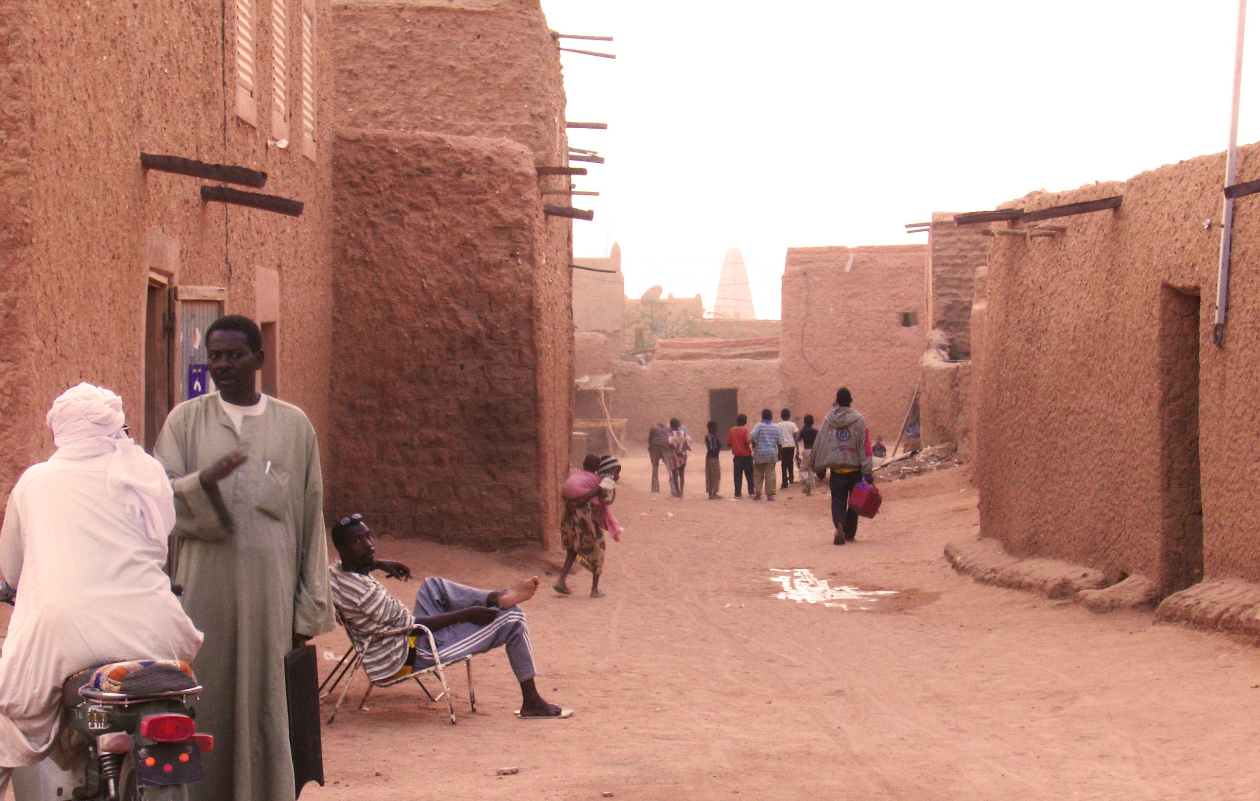 L'image montre une rue dans un village traditionnel, probablement situé dans une région désertique. On y voit des maisons en terre, avec des murs de couleur ocre. Plusieurs personnes déambulent tranquillement dans la rue, certaines s'arrêtent pour discuter. À gauche, un homme est assis sur une moto, tandis qu'un autre se repose sur une chaise. L'atmosphère est calme et chaleureuse, avec un éclairage doux peut-être dû au coucher du soleil. Les ombres et la poussière soulignent le caractère rustique du lieu.