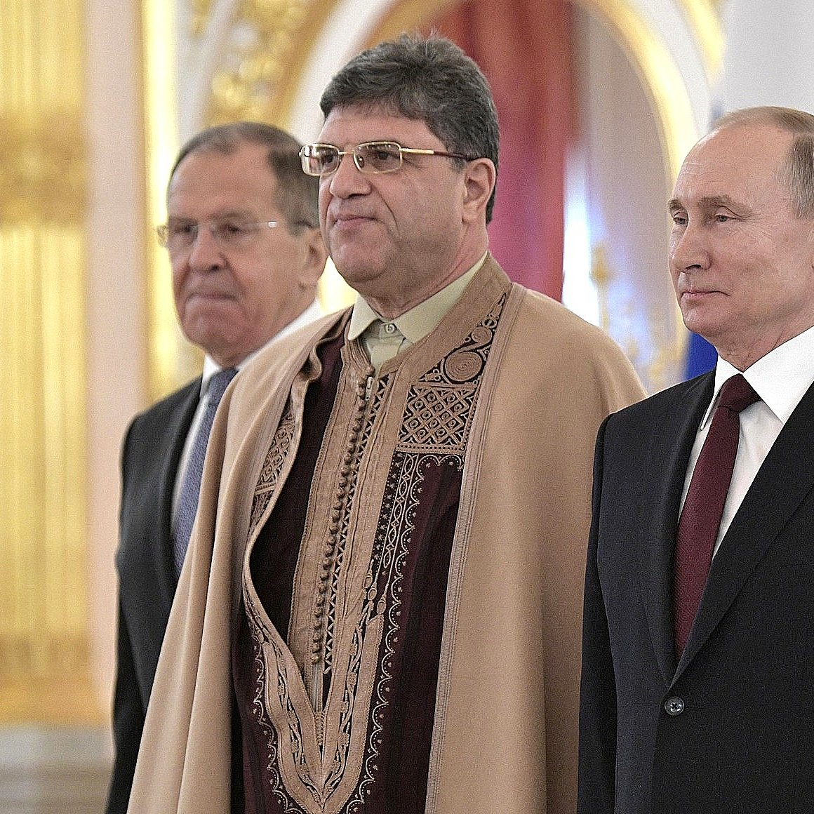 در این تصویر، سه نفر در یک مراسم ایستاده‌اند. فرد سمت راست ولادیمیر پوتین، رئیس‌جمهور روسیه، است که یک کت رسمی به تن دارد. فرد وسط، ظاهری متفاوت با لباس سنتی دارد و در حالتی آرام ایستاده است. فرد سمت چپ نیز لباسی رسمی به تن دارد و به سمت جلو نگاه می‌کند. پس‌زمینه تصویر شامل جزئیات مجلل و تزئینات داخلی است که نشان‌دهنده فضایی رسمی و تشریفاتی است.