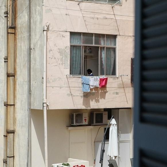 L'image montre un immeuble avec des murs de couleur rose. À un des balcons, on aperçoit un enfant qui semble regarder à l'extérieur. Des vêtements sont étendus sur la rambarde du balcon, et au sol, on peut voir un parasol ouvert. À droite, un volet est partiellement ouvert, ajoutant une autre dimension à la scène. L'ambiance semble calme et typique d'un environnement urbain.