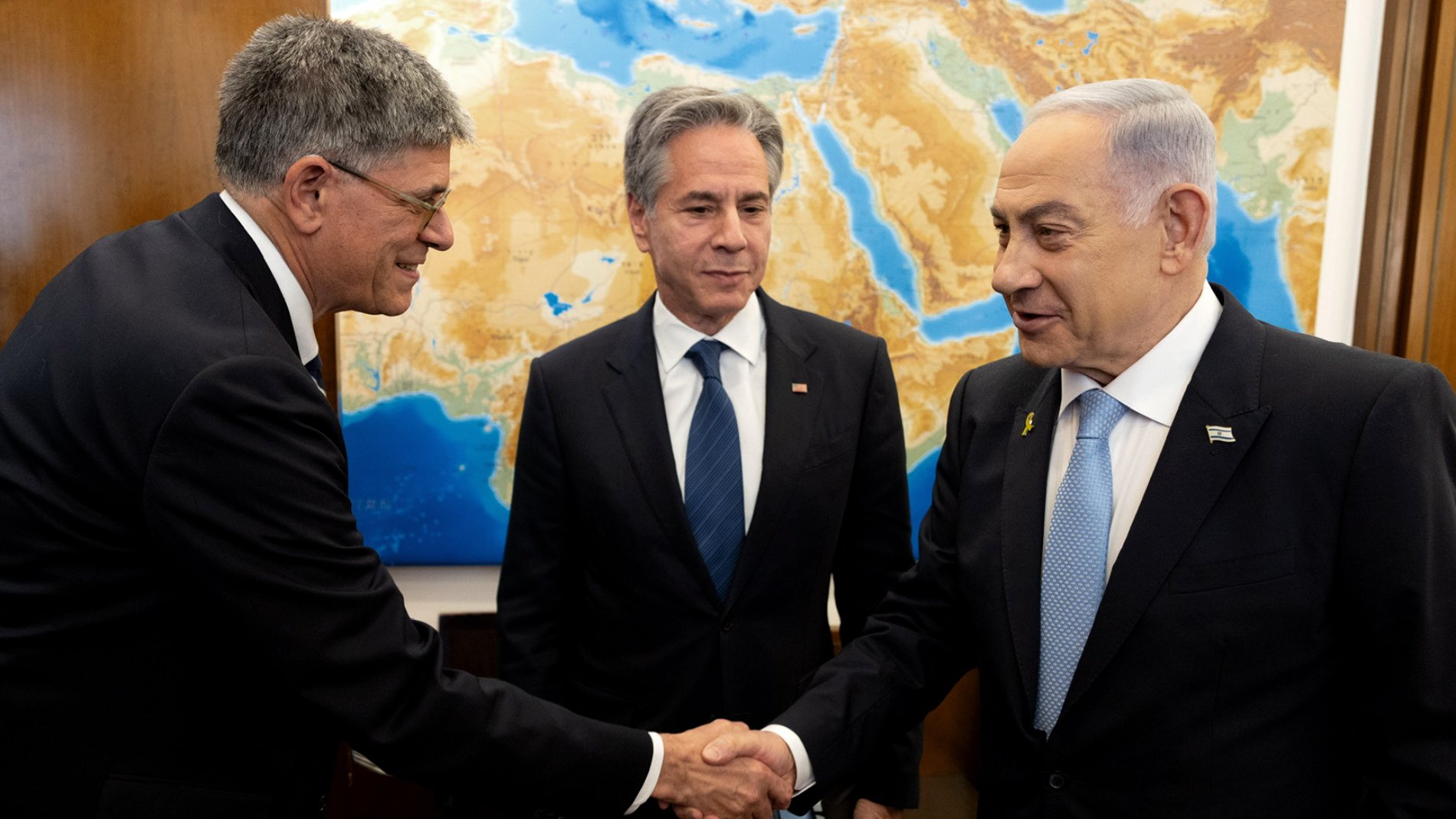 در تصویر، چند نفر در حال گپ‌وگفت هستند. دو نفر با هم دست می‌دهند و یکی دیگر در پس‌زمینه ایستاده است. پس‌زمینه شامل نقشه‌ای از منطقه خاورمیانه است. افراد در حال گفتگو به نظر می‌رسند و حالت چهره آن‌ها نشان‌دهنده تعامل مثبت است. لباس‌های رسمی آنها به اهمیت جلسه اشاره دارد.