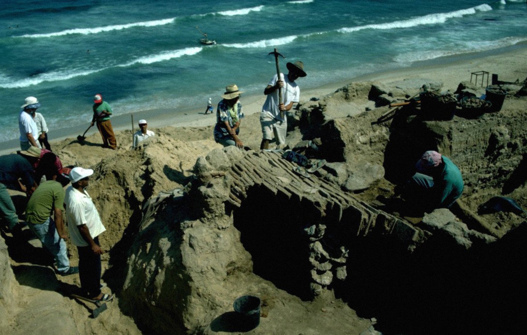 A group of people excavates a sandy site near the ocean, surrounded by waves and tools.