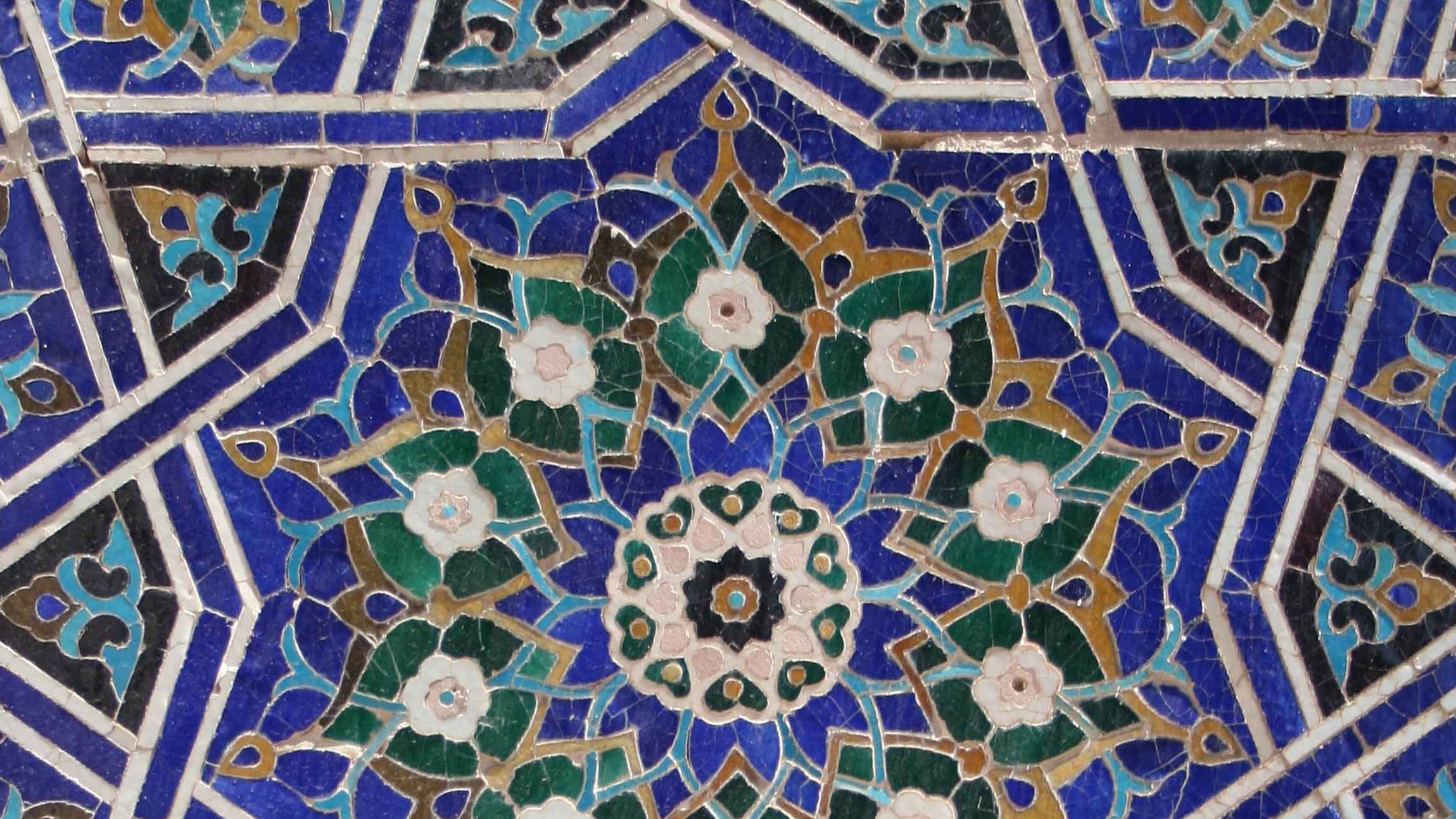 تظهر الصورة زخارف معمارية دقيقة، تتكون من أنماط هندسية وأزهار بألوان متنوعة مثل الأزرق والأخضر والكريمي. التصميم يبرز تداخل الأشكال بشكل متناظر، مع تفاصيل دقيقة تضيف جمالًا وتناسقًا للمظهر. يمكن رؤية تأثير الفنون الإسلامية التقليدية في هذا العمل، مما يعكس مهارة الحرفيين ودقتهم في تنفيذ مثل هذه الزخارف.