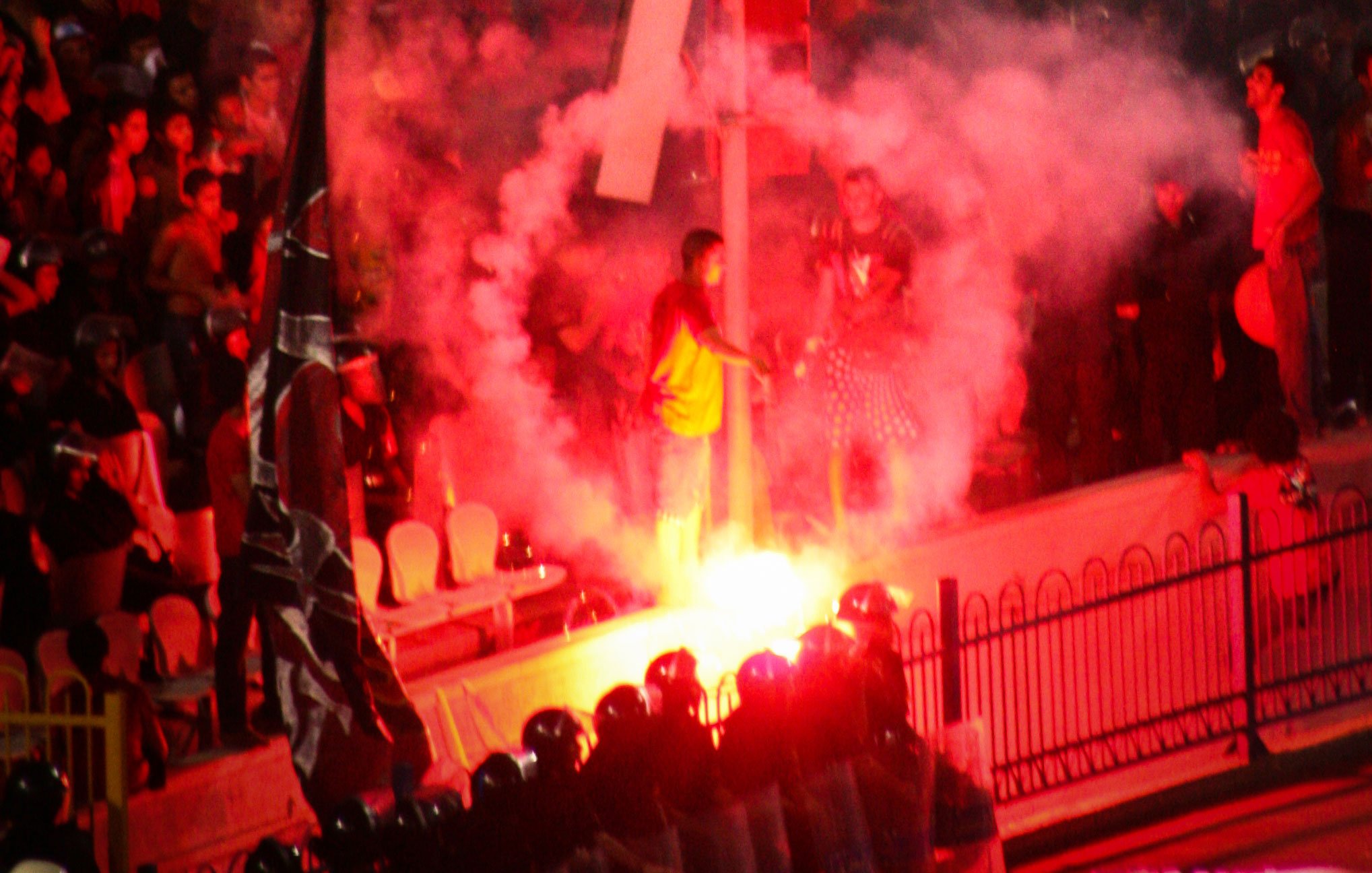 L'image montre une scène de manifestation avec une atmosphère intense. On peut voir des fumigènes rouges créant une nuée de fumée, ainsi que des personnes rassemblées autour d'une structure ou d'un symbole marquant, probablement dans un contexte de protestation. Au premier plan, une file de policiers en uniformes semble faire face aux manifestants. L'ensemble de la scène transmet une impression de tension et d'engagement émotionnel.