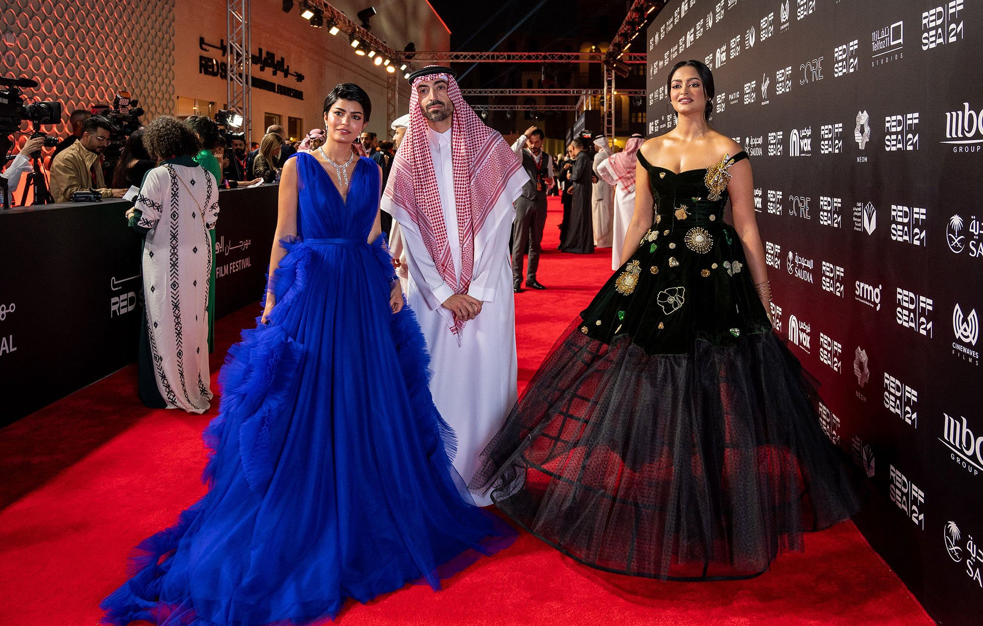 L'image montre trois personnes posant sur un tapis rouge lors d'un événement. À gauche, une femme porte une robe de soirée bleue élégante avec des volants. Au centre, un homme habillé en tenue traditionnelle, comprenant un thobe et un chapeau saoudien. À droite, une femme porte une robe noire ornée de motifs floraux, ajoutant une touche sophistiquée à son look. L'arrière-plan montre un décor stylisé, typique des événements glamour.