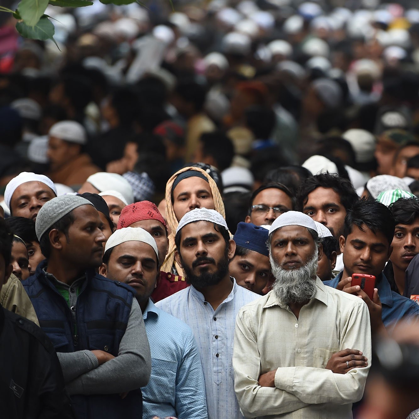 این تصویر نشان‌دهنده جمعیتی بزرگ از مردان است که در مکانی شلوغ تجمع کرده‌اند. بسیاری از افراد کلاه‌های سنتی اسلامی یا «تاج» به سر دارند و در حال انتظار یا نظارت بر چیزی هستند. چهره‌ها متفاوت‌اند و برخی از افراد با لباس‌های ساده و دیگران با لباس‌های رسمی‌تر مشخص شده‌اند. جو کلی تصویر احساس تمرکز و انتظار را منتقل می‌کند.