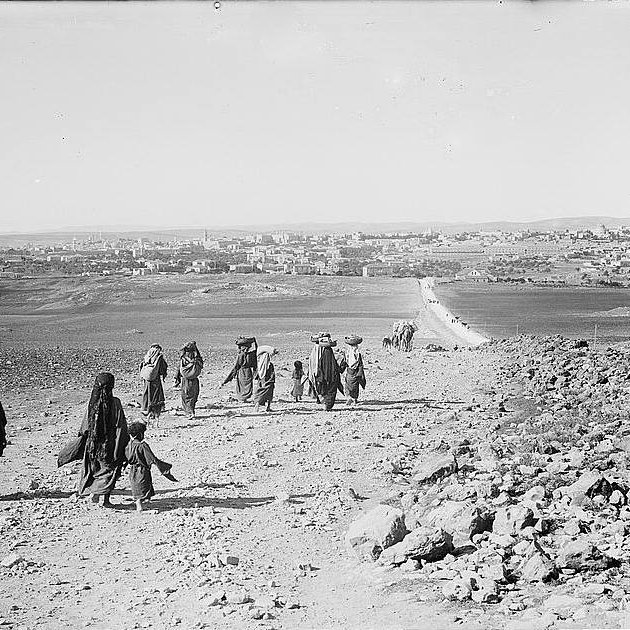 The image depicts a group of people walking along a rocky path in a barren landscape. They appear to be headed toward a distant town or village, which is visible in the background. The group consists of several adults, some of whom are carrying burdens on their heads, and a child walking alongside them. The terrain is dry and rugged, with scattered rocks and sparse vegetation. The sky is clear, and the scene conveys a sense of movement and migration through a rural setting.
