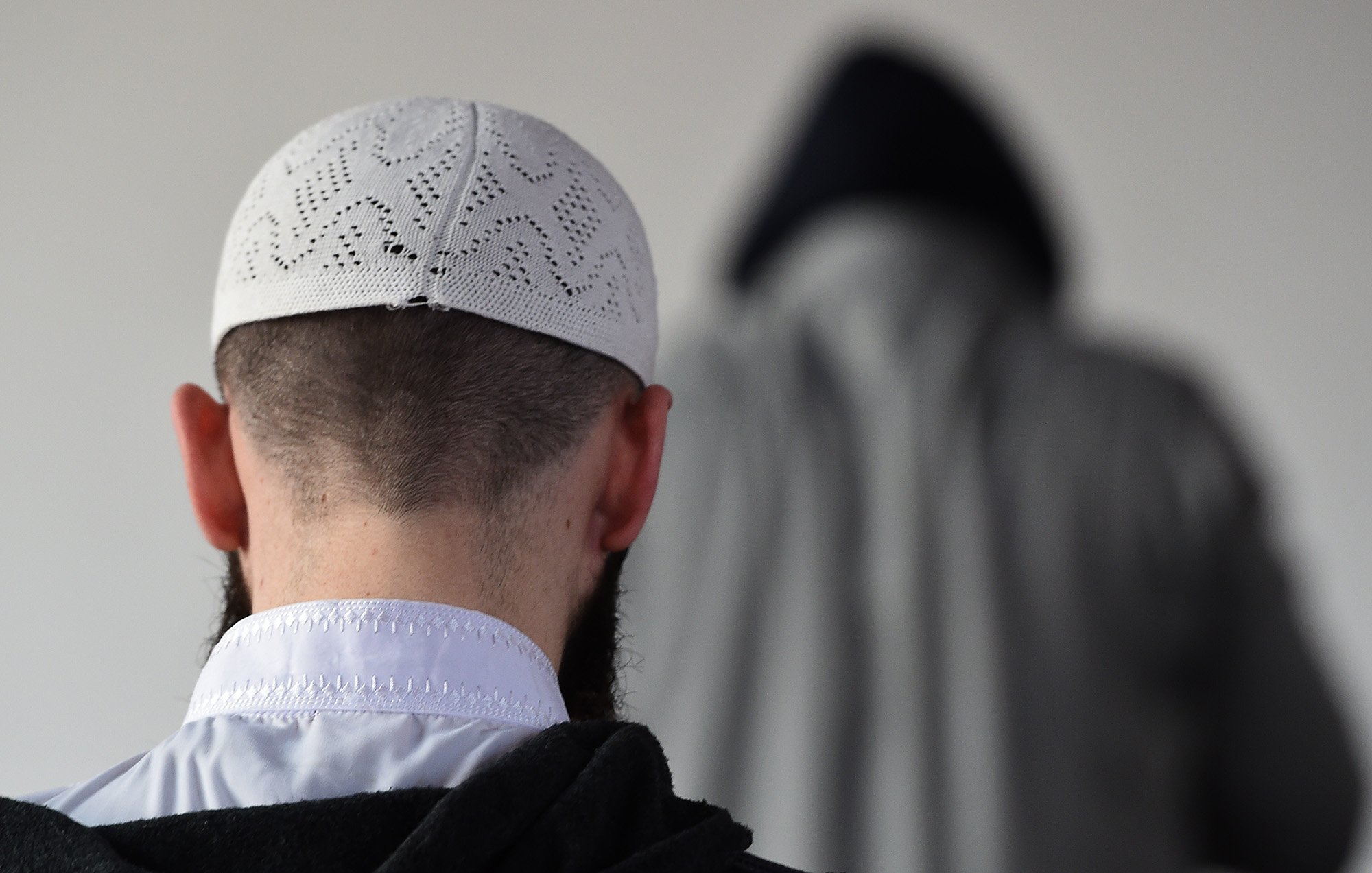 الصورة تظهر شخصًا يرتدي قبعة تقليدية بيضاء، مع ظهور جزء من رأسه وظهره. خلفه يوجد شخص آخر يرتدي غطاء رأس داكن مع معطف، مما يخلق تباينًا في الألوان. الجو العام للصورة يوحي بالهدوء والتركيز، وقد تكون في سياق ديني أو روحي.