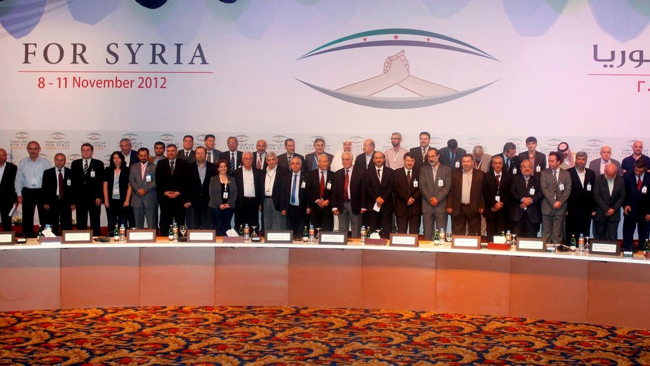 L'image montre un groupe de personnes réunies lors d'un événement formel, probablement une conférence. Elles sont alignées en plusieurs rangées, toutes en costume, et semblent participer à un rassemblement lié à la Syrie, comme indiqué par le texte en arrière-plan. Le décor est coloré, avec des drapeaux et des motifs qui évoquent l'événement. Les participants adoptent des expressions sérieuses, ce qui suggère l'importance de l'occasion.