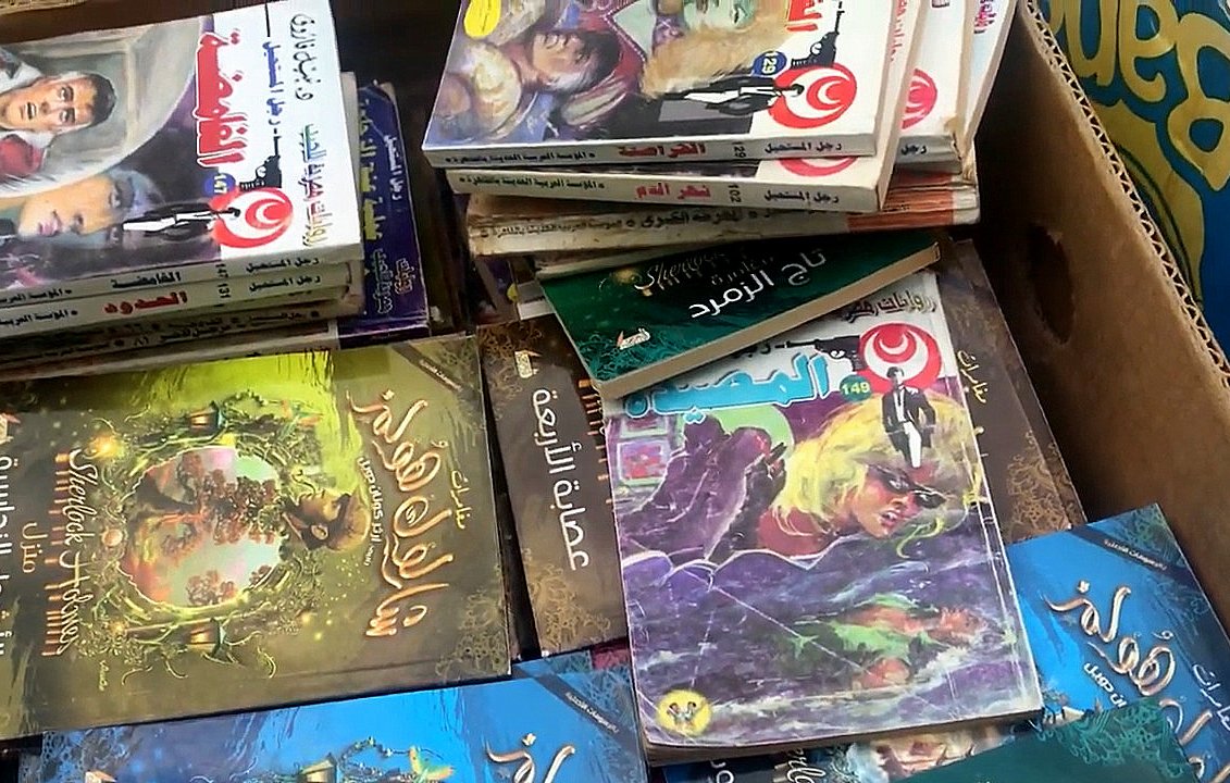 L'image montre une boîte remplie de livres empilés les uns sur les autres. On peut apercevoir différents titres en arabe sur les couvertures, illustrés par des images colorées. Certains livres semblent appartenir à des séries de contes ou de romans graphiques, avec des illustrations captivantes et des textes en arabe. L'ensemble donne une impression d'abondance littéraire.