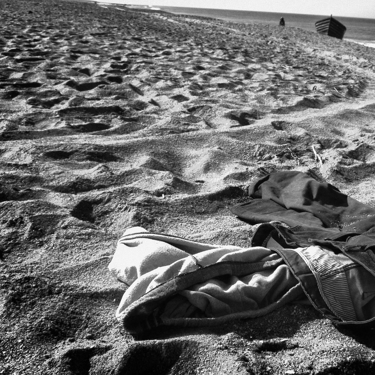 تظهر في الصورة ملابس متكدسة على الرمال على شاطئ. يبدو أن الشاطئ هادئ، حيث تُظهر الأمواج البعيدة خط الأفق. الرمال تحمل آثار أقدام وكأن هناك نشاطًا قد حدث في المنطقة. الأجواء بشكل عام تعكس شعورًا بالوحدة والهدوء.