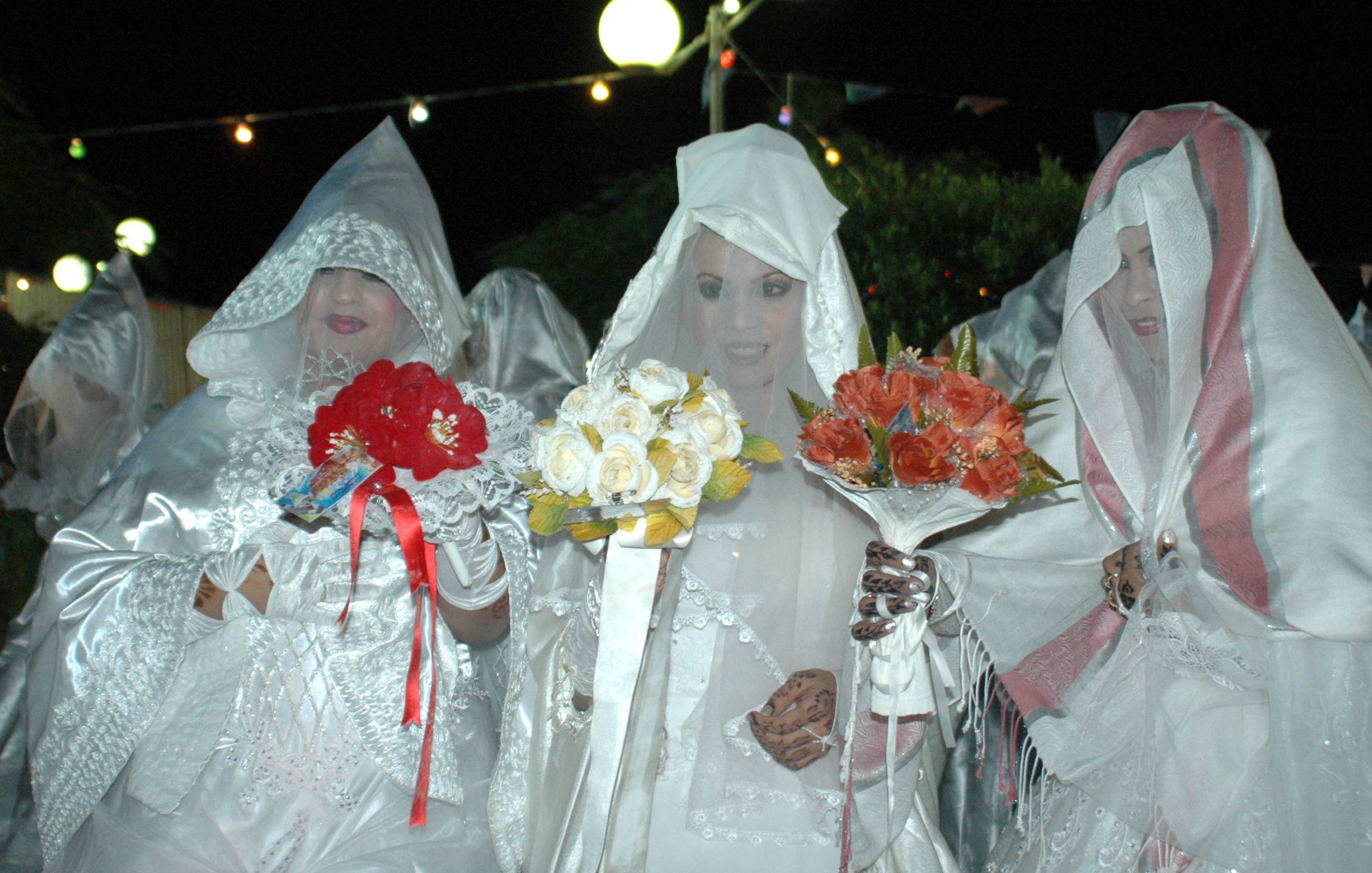 The image features three women dressed in elaborate white outfits that resemble bridal attire. They are wearing head coverings and are holding bouquets of flowers. The setting appears to be outdoors at night, illuminated by soft lights above them. The overall atmosphere seems festive, possibly related to a celebration or cultural event. The women's faces are mostly obscured, adding a mysterious or dramatic effect to the scene.