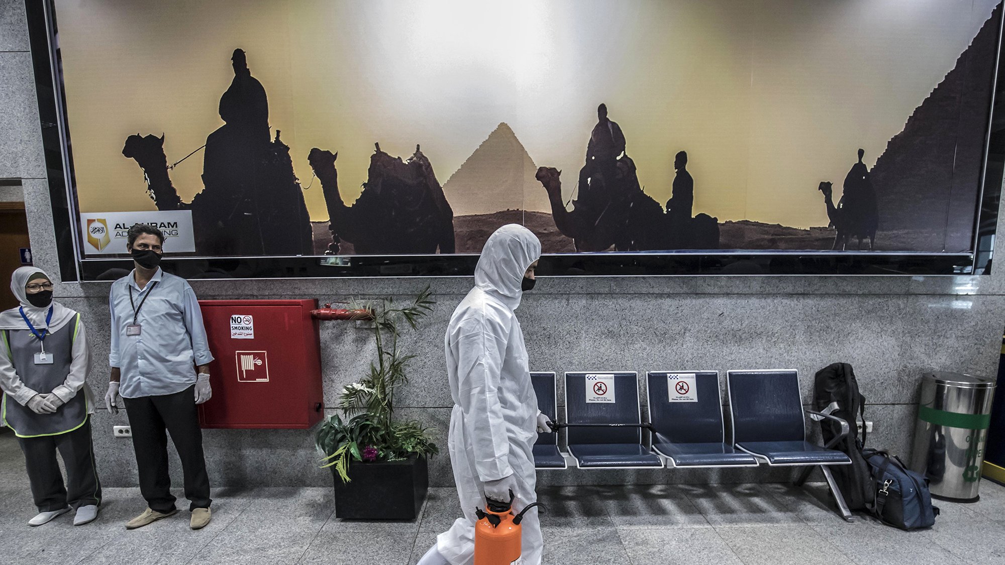 در این تصویر، یک فرد در حال قدم زدن است که لباسProtective پوشیده و یک جعبه نارنجی رنگ در دست دارد. در پس‌زمینه، تصویری از اهرام مصر و افرادی که بر روی شترها نشسته‌اند، به صورت سیاه و سفید دیده می‌شود. همچنین، دو نفر دیگر در سمت چپ تصویر ایستاده‌اند و ماسک زده‌اند. فضای کلی تصویر گویا محیطی عمومی یا فرودگاهی است.