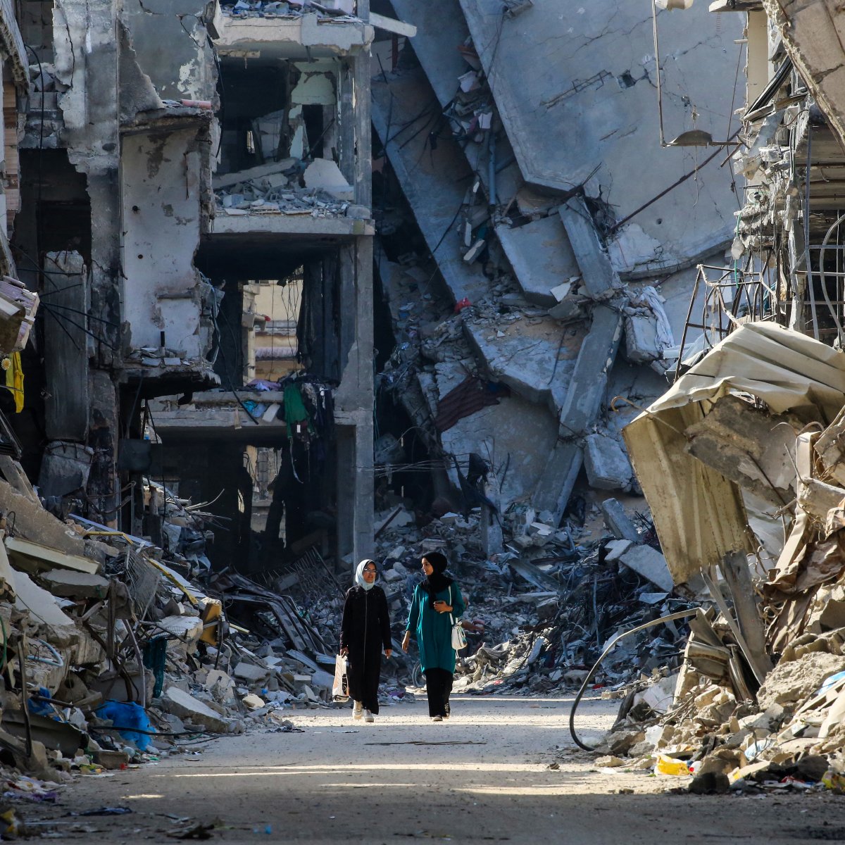 L'immagine mostra due persone che camminano in una strada distrutta da un conflitto, circondate da edifici in rovina. Le strutture sono parzialmente crollate e si vedono macerie sparse ovunque, creando un'atmosfera di desolazione e devastazione. La scena evidenzia la gravità della situazione, con un forte contrasto tra le persone che camminano e l'ambiente circostante, che riflette la difficoltà e la sofferenza di un'area colpita dalla guerra.