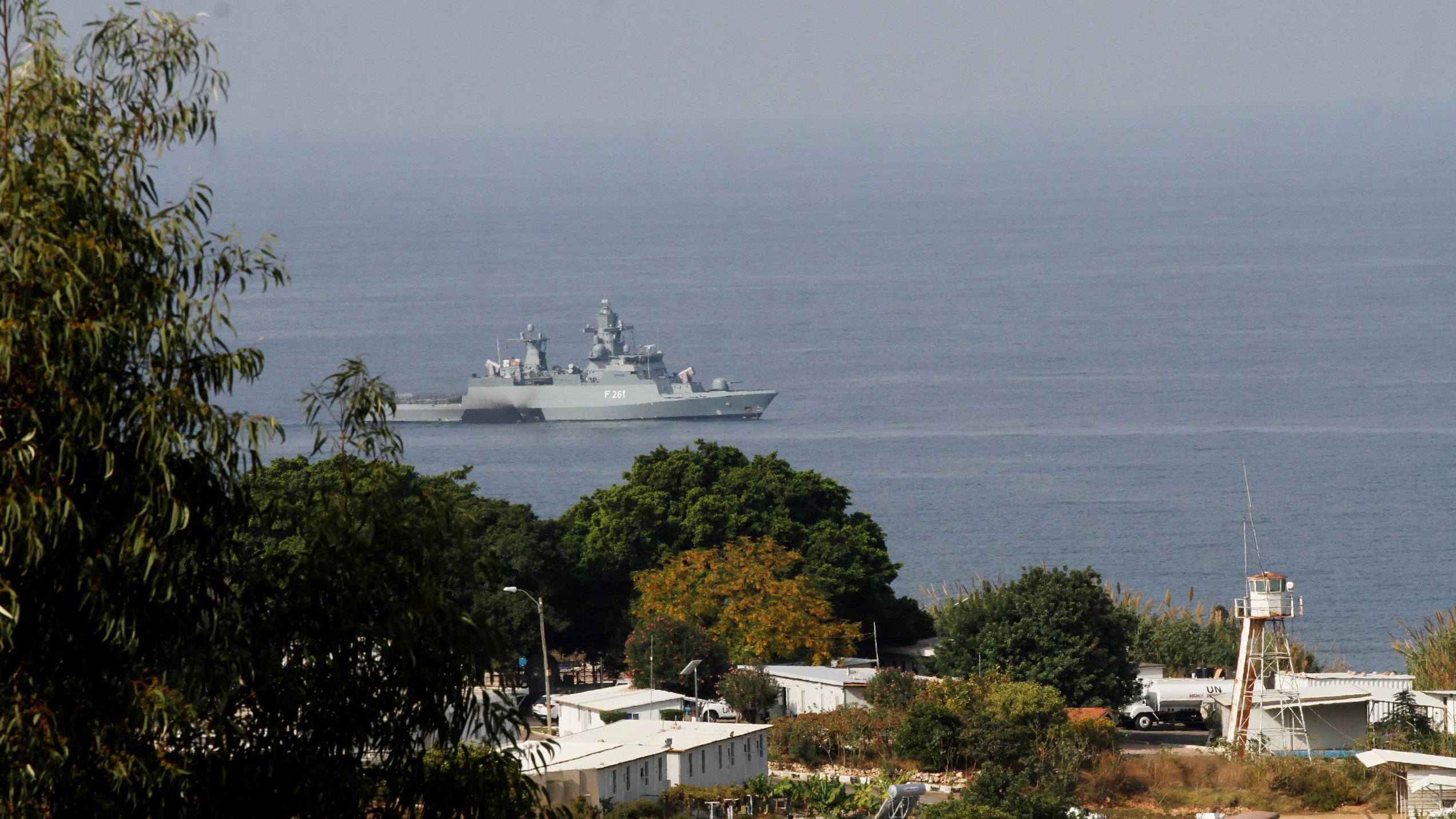 تظهر في الصورة سفينة حربية تجوب المياه بالقرب من ساحل يطل على منطقة خضراء. في الخلفية، يمكن رؤية بعض المباني والمرافق، كما يُلاحظ وجود برج مراقبة. السماء تبدو صافية، والمشهد يعكس أجواء بحرية هادئة.