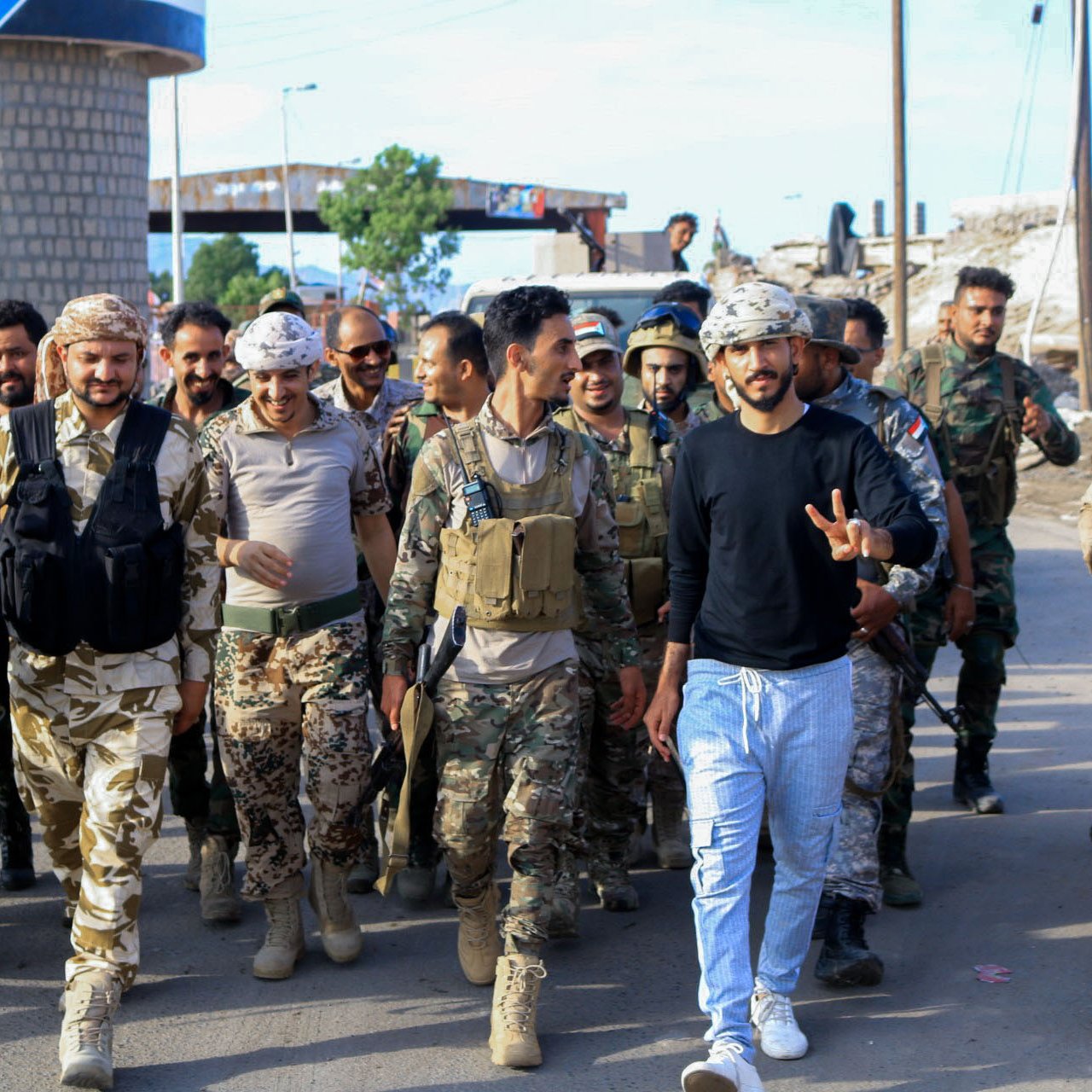 L'image montre un groupe de soldats marchant ensemble dans une zone urbaine. Parmi eux, on peut voir des militaires en uniformes camouflés et d'autres en tenues militaires variées. Certains portent des gilets tactiques et des casques. On remarque une atmosphère de camaraderie et de détermination parmi les membres du groupe, avec une personne au centre faisant un signe de paix. En arrière-plan, des infrastructures urbaines sont visibles, ce qui indique un environnement de mission militaire.
