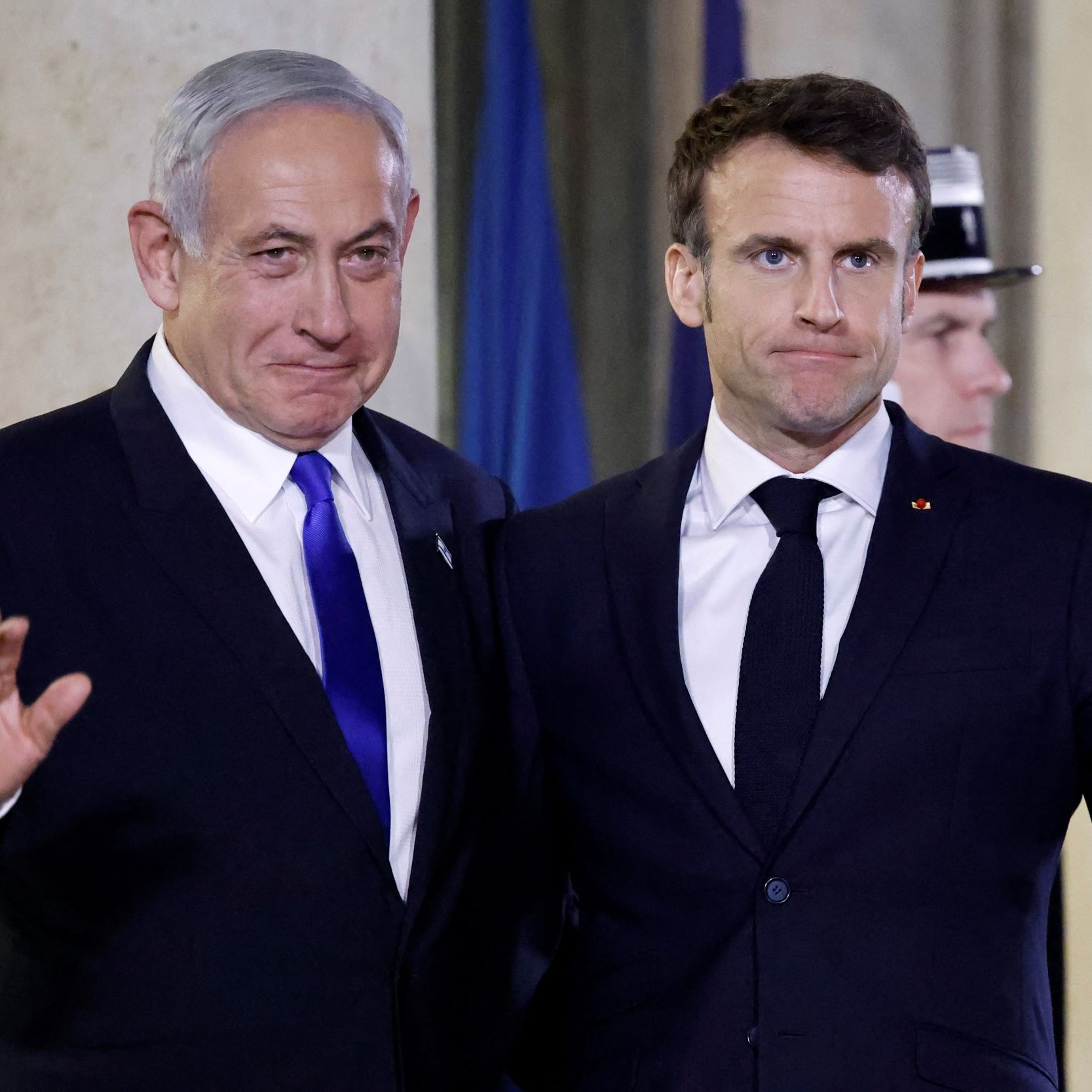 The image shows two men standing side by side, both dressed in formal attire. One is wearing a blue tie and the other has a dark tie, indicating a formal event. They appear to be waving, possibly to an audience or camera. The background suggests an official setting, likely an indoor venue with flags or formal decorations. Their expressions seem to convey a mix of seriousness and cordiality.