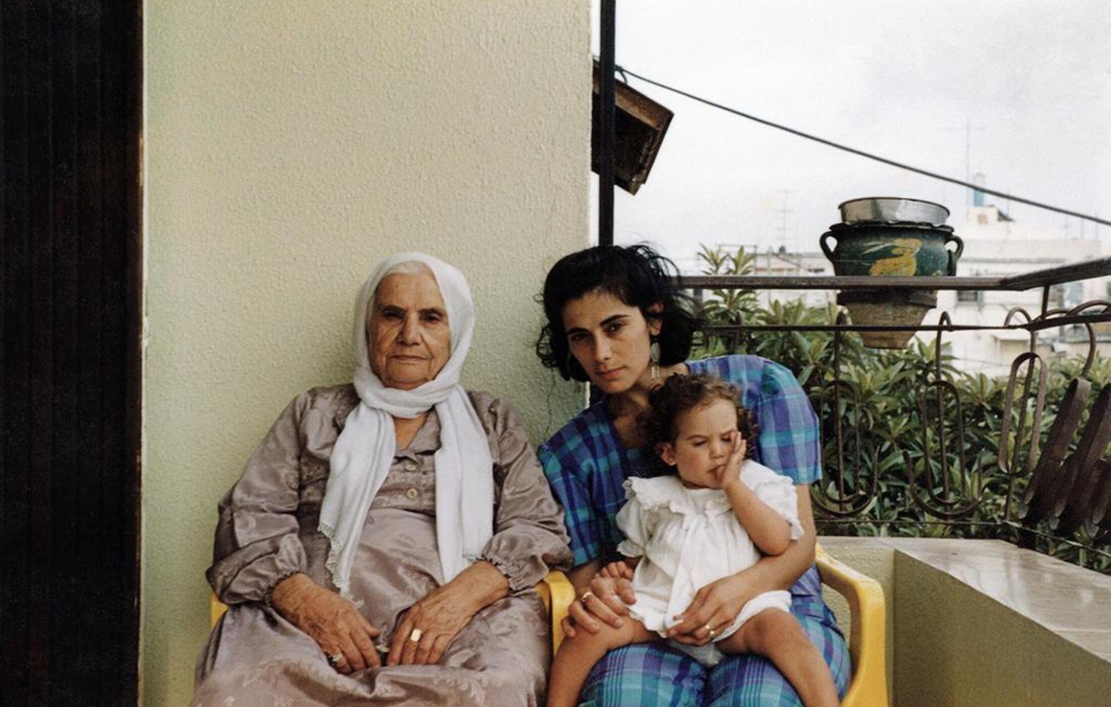 L'image montre trois femmes assises sur un balcon. À gauche, une femme âgée porte une robe grise et un foulard blanc. Au centre, une jeune femme, avec les cheveux noirs et bouclés, regarde vers l'objectif tout en tenant un enfant. L'enfant, assis sur les genoux de la jeune femme, semble jouer avec quelque chose. Le fond montre un environnement urbain avec des plantes et des éléments architecturaux. L'ambiance de l'image est chaleureuse et familiale.