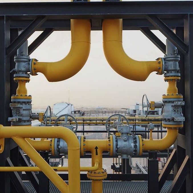 La imagen muestra una estructura industrial, probablemente de una planta de procesamiento de petróleo o gas. Hay varias tuberías de color amarillo que se conectan entre sí, junto con válvulas y otros elementos metálicos. El fondo sugiere un paisaje industrial con tanques y equipos, lo que indica un entorno de trabajo relacionado con la energía o la petroquímica. La escena parece ser al aire libre, bajo un cielo claro.