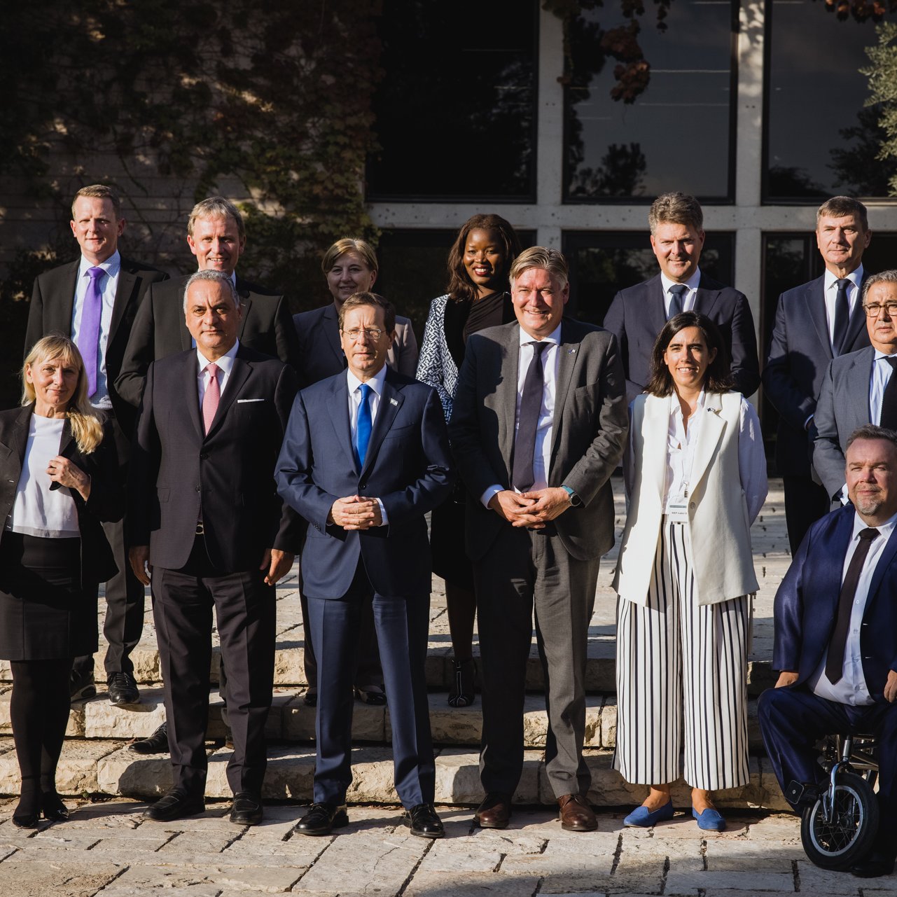 الصورة تظهر مجموعة من الأشخاص يقفون معًا في بيئة خارجية، ربما تكون حديقة أو منطقة أمامية لمبنى. يوجد في الخلفية مشهد طبيعي مع أشجار ونباتات. يرتدي الحضور ملابس رسمية وأنيقة، مما يشير إلى أن الصورة قد تم التقاطها في مناسبة رسمية أو مؤتمر. تتراوح تعبيرات الوجوه بين الابتسامات والجدية، مما يعكس جوًا من التعاون والتواصل.