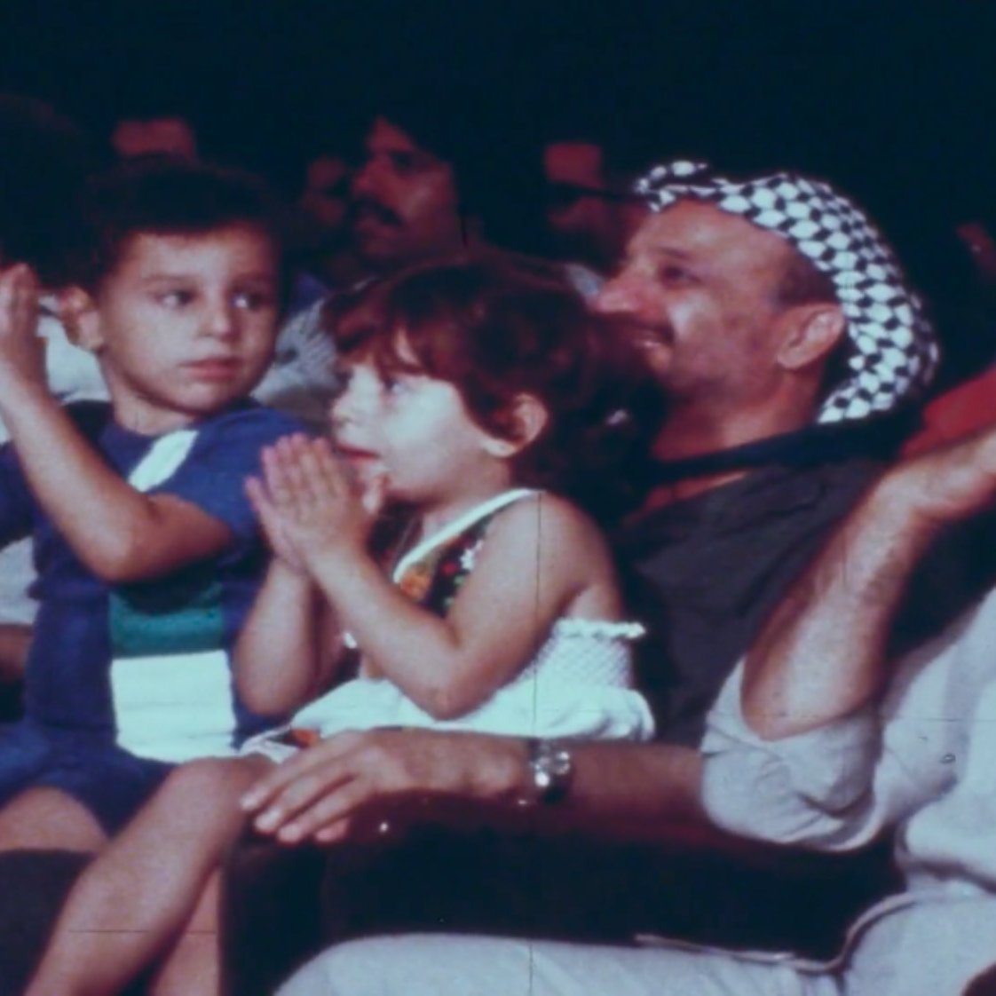 La imagen muestra un grupo de personas sentadas en un evento, aplaudiendo. En el centro de la imagen, hay dos niños: un niño que lleva una camiseta de colores y una niña que lleva un vestido claro. Detrás de ellos, se puede ver a un hombre con una keffiyeh (un pañuelo típicamente árabe) y otros asistentes en el fondo. La atmósfera parece festiva, y todos están disfrutando del momento.