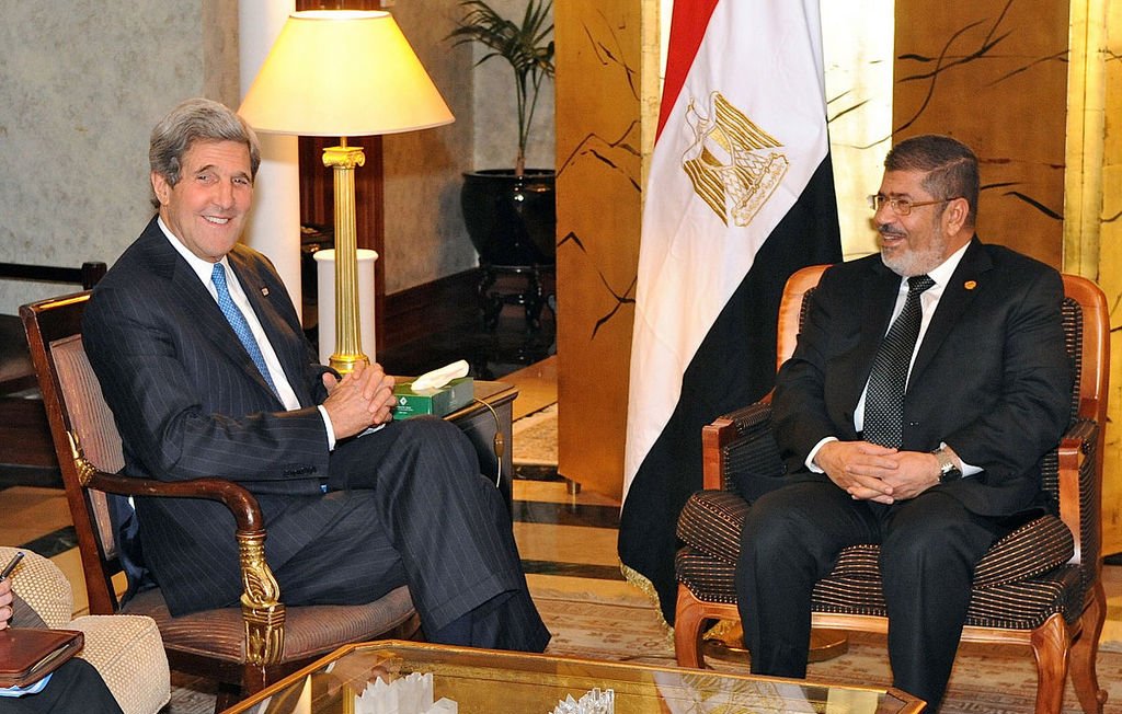 تظهر الصورة اجتماعًا بين شخصين يجلسان في غرفة مريحة. يجلس أحدهما على اليسار وهو يرتدي بدلة رسمية مع ربطة عنق ويبتسم، بينما يجلس الشخص الآخر على اليمين، كذلك يرتدي بدلة ويبدو سعيدًا. الخلفية تحتوي على علم مصر، مما يشير إلى أن الاجتماع قد يكون ذو طابع رسمي يتعلق بالشؤون السياسية أو العلاقات الدولية. الأجواء تبدو ودية وإيجابية.