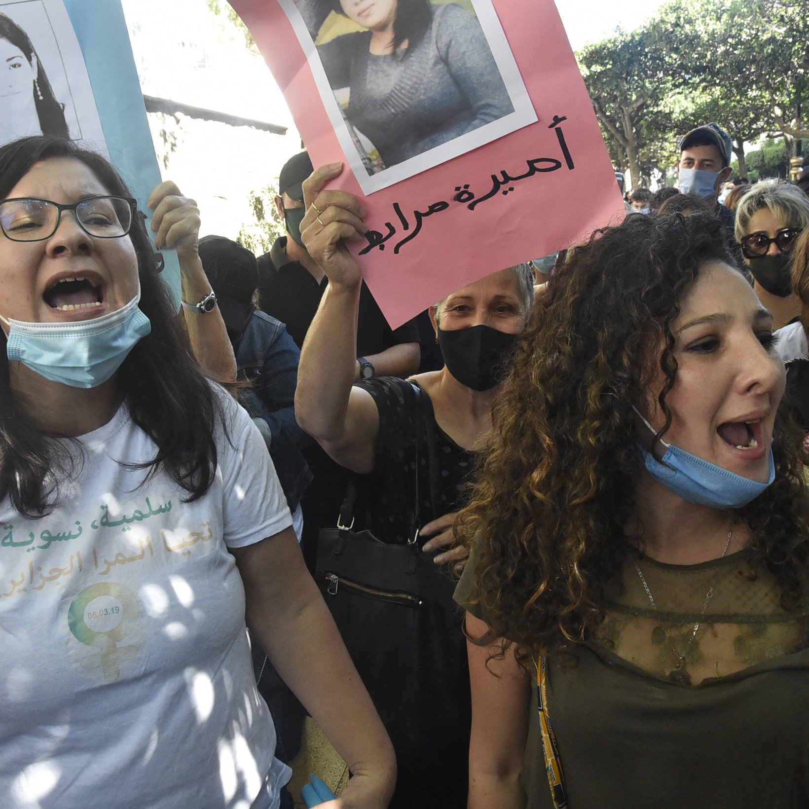 En la imagen se observa a un grupo de personas participando en una protesta. Varias mujeres están al frente, gritando con energía y sosteniendo carteles que muestran fotografías de otras personas. Algunas de las asistentes llevan mascarillas. El ambiente parece lleno de emoción y determinación, con un fondo de árboles y una calle. Las pancartas tienen inscripciones en diferentes idiomas, lo que sugiere que se trata de una manifestación con un propósito importante.