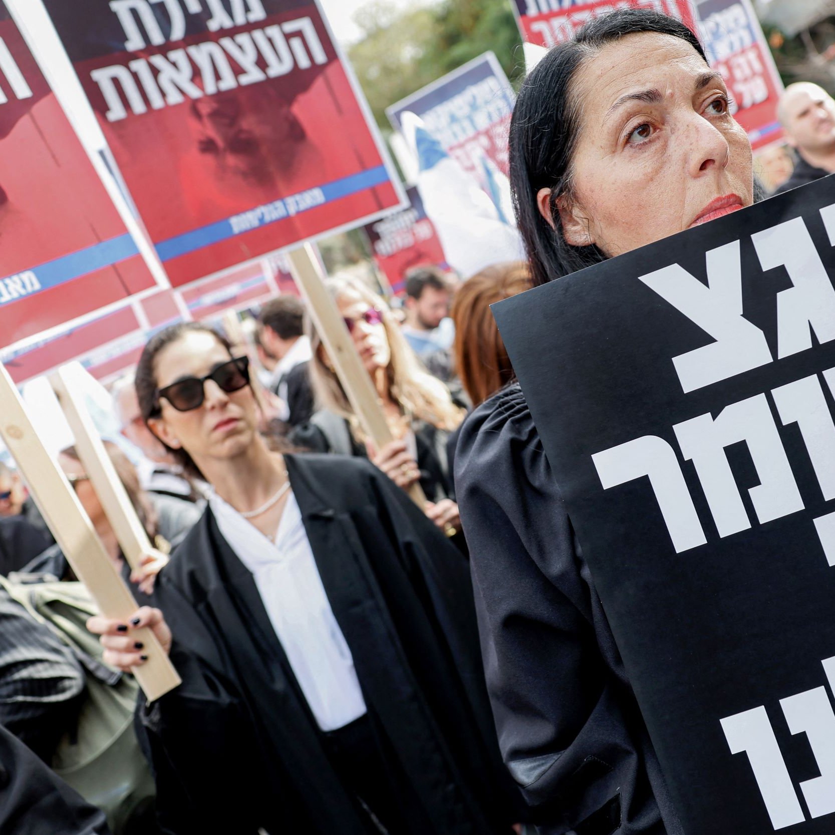 تظهر الصورة تجمعًا حاشدًا من المحتجين، حيث يرتدي المشاركون ملابس تشبه الأرواب القانونية. تركز الكاميرا على امرأة تحمل لافتة كبيرة مكتوب عليها نص بخط عريض. خلفها، يمكن رؤية آخرين يحملون لافتات أيضًا. يبدو أن المحتجين يعبرون عن مطالب معينة أو يعبرون عن احتجاجهم إزاء قضية ما. تعبر تعابير وجه المشاركين عن الجدّية والاهتمام بالقضية المطروحة.