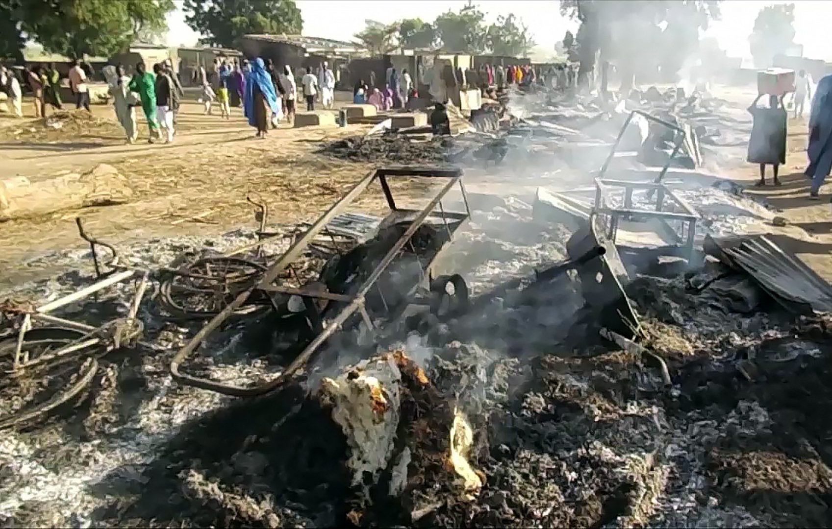 La imagen muestra un escenario devastador después de un incendio. Hay estructuras de metal chamuscadas y escombros humeantes en el suelo. En el fondo, algunas personas parecen estar observando la escena, mientras que el humo se eleva en el aire. El paisaje refleja una atmósfera de destrucción, posiblemente en un área rural o un mercado.