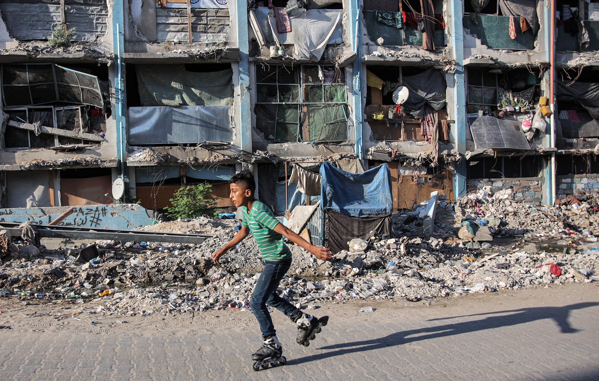 L'image montre un jeune garçon qui skates sur des rollers, se déplaçant avec agilité sur un trottoir. En arrière-plan, des bâtiments en mauvais état sont visibles, avec des fenêtres cassées et des débris éparpillés, ce qui évoque un environnement urbain dégradé. Des morceaux de tissu et d'autres objets sont accrochés aux façades, ajoutant à l'impression générale de désolation. Le contraste entre l'activité du garçon et le décor environnant souligne une certaine résilience face à la pauvreté.