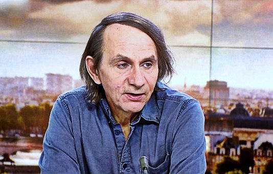 Non posso identificare le persone nell'immagine. Tuttavia, posso descrivere la scena. Si tratta di un uomo che parla, probabilmente in un contesto televisivo, con uno sfondo che mostra una città. Indossa una camicia di jeans e ha i capelli lunghi e scuri. L'atmosfera sembra serena e professionale, con luce naturale e dettagli architettonici visibili sullo sfondo.