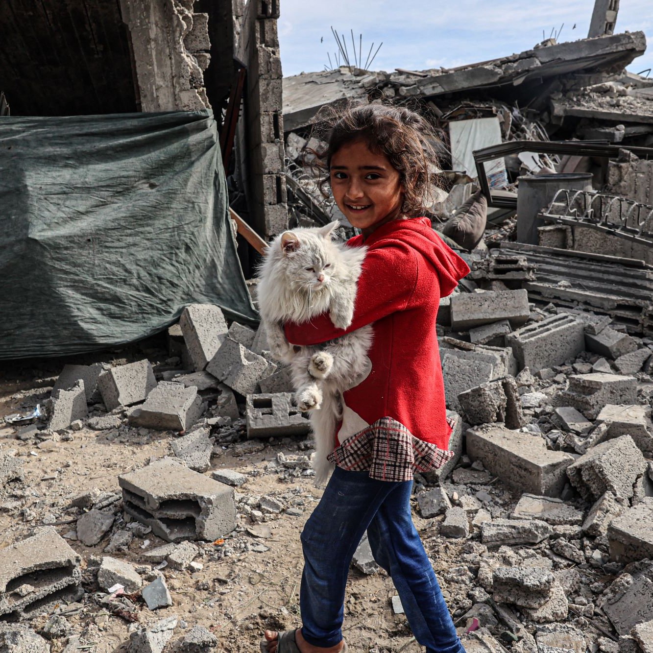 L'immagine mostra una bambina che sorride mentre tiene in braccio un coniglietto bianco. Si trova in un ambiente distrutto, con macerie e rovine visibili intorno a lei. Il suo volto esprime gioia nonostante il contesto difficile, e indossa un maglione rosso e jeans. Sullo sfondo si vedono resti di edifici e teli di plastica, che evidenziano una situazione di emergenza.
