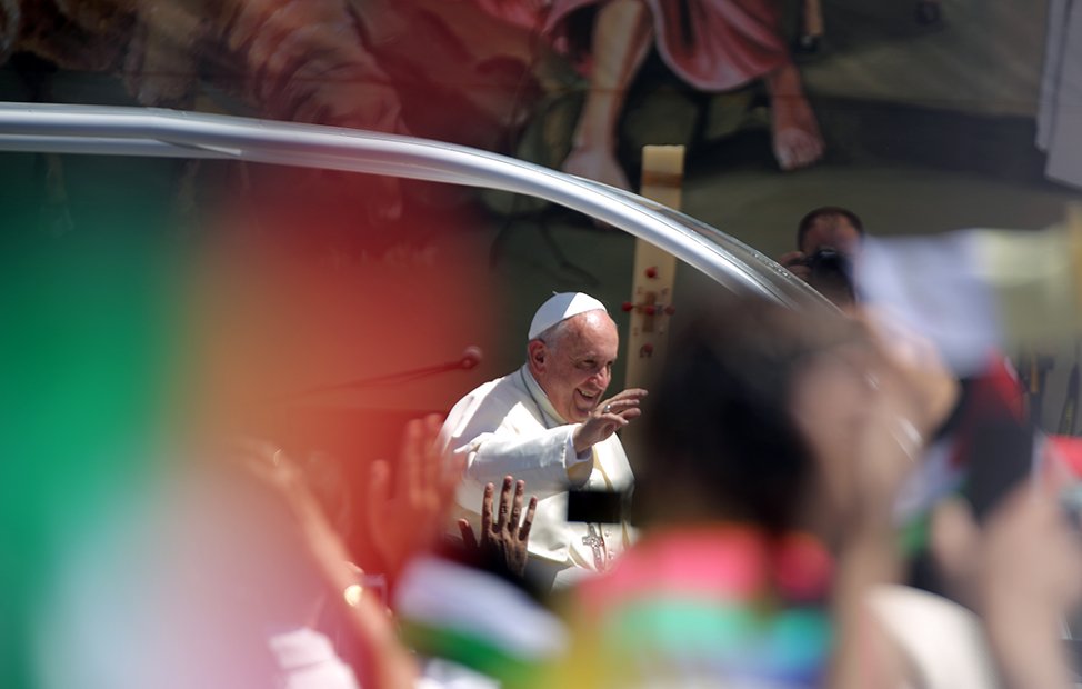 L'image montre une scène d'un événement public avec le Pape François. Il est souriant et fait un geste de la main en saluant la foule. On peut apercevoir des drapeaux multicolores dans le premier plan, ce qui indique une ambiance festive et dynamique. En arrière-plan, on distingue des illustrations ou des représentations qui semblent évoquer une célébration ou un rassemblement spirituel. L'ensemble dégage une atmosphère de joie et d'enthousiasme de la part des personnes présentes.