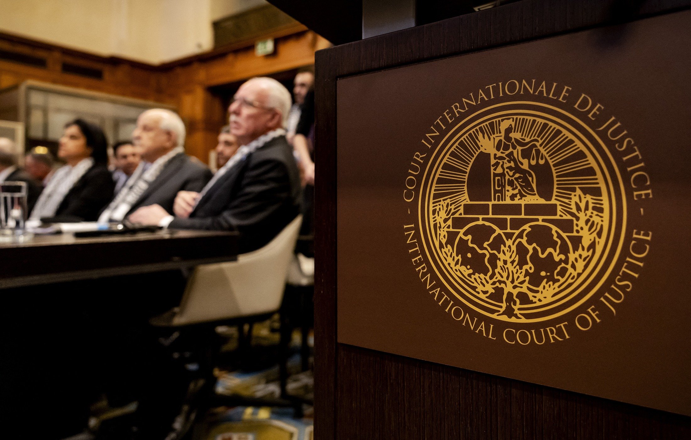 تظهر الصورة مشهدًا من داخل قاعة محكمة، حيث يجلس مجموعة من الأشخاص في الخلفية، ويبدو أنهم يستمعون لمداولات أو حديث مهم. في المقدمة، يوجد شعار المحكمة الدولية للعدل، وهو يتضمن تصميمًا معقدًا يرمز إلى العدالة. الطراز المعماري للقاعة يظهر الفخامة والرسمية. الأجواء تعكس أهمية الحدث الحاصل داخل هذه المحكمة.