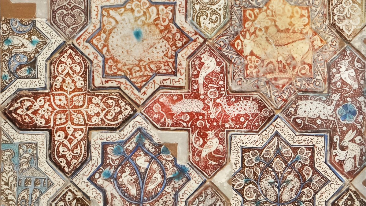 L'image présente un motif complexe et orné, typique de l'art islamique. On y voit des formes géométriques et des étoiles, réalisées dans des teintes richement variées allant des rouges aux bruns, avec des touches de bleu et de blanc. Les motifs sont décorés de détails floraux et d'animaux stylisés, ajoutant à la richesse visuelle de l'ensemble. Cette composition évoque une harmonie artistique ainsi qu'un savoir-faire traditionnel, caractéristique des mosaïques ou des carreaux décoratifs.
